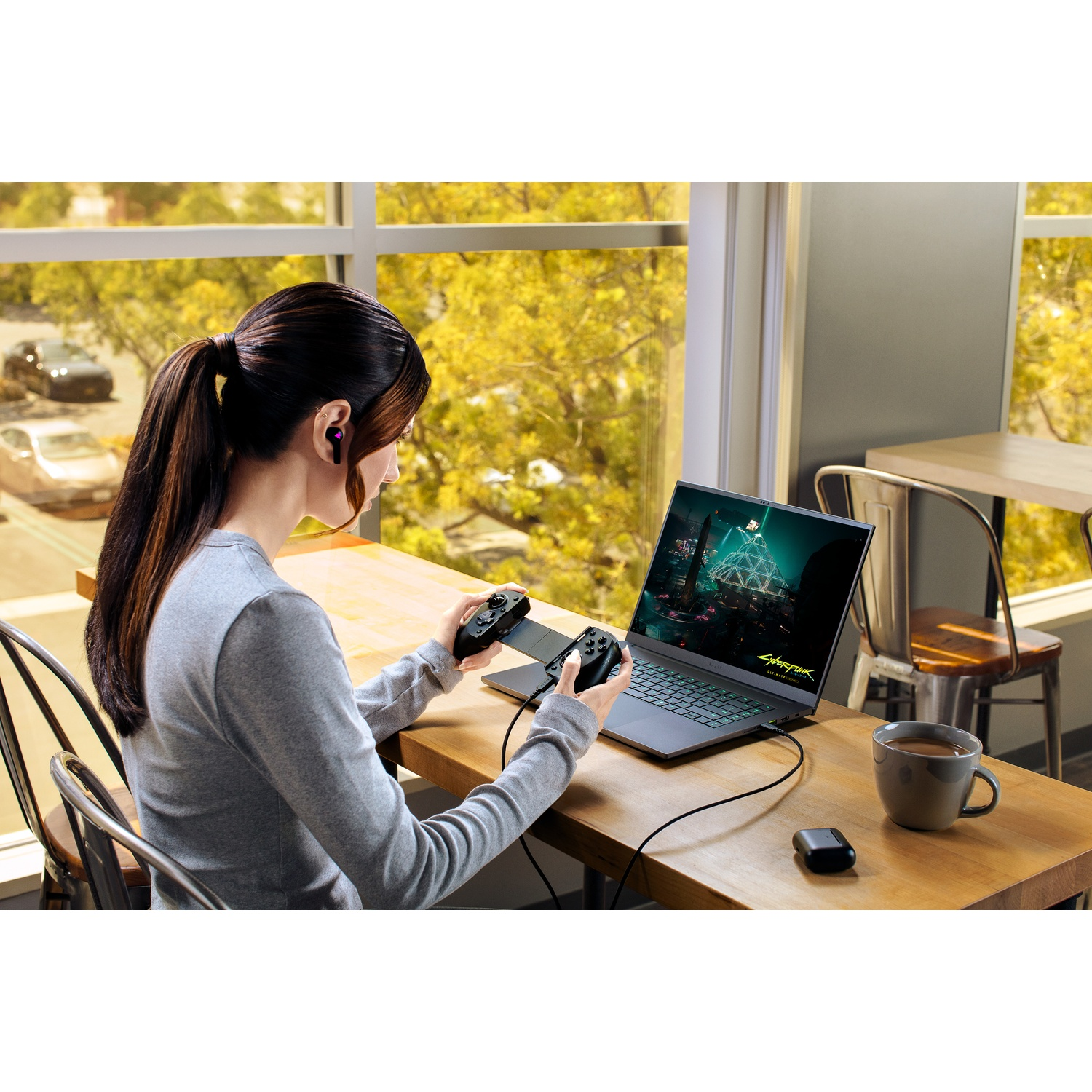 Vrouw gamet op laptop, controllers vasthoudend. Scherm toont Cyberpunk. Koffie, oortelefoons en auto's buiten zichtbaar.