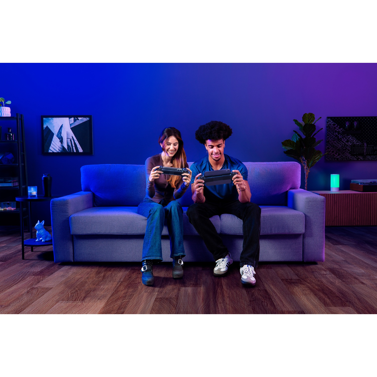 Two people are sitting on a couch, holding game controllers. They are smiling. The background is a blue wall with a plant and decor.