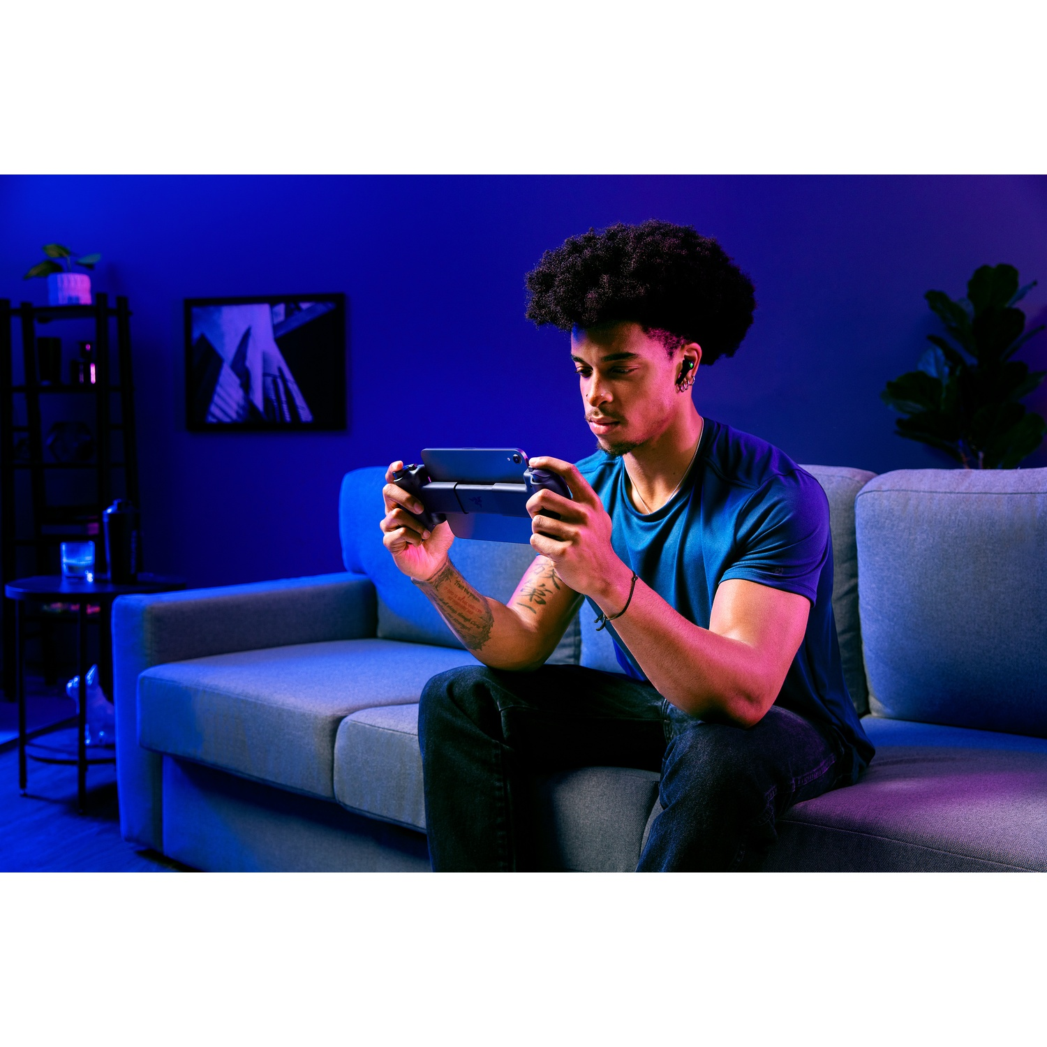 A man with headphones plays a handheld game console, sitting on a gray couch. The room has blue and purple lighting.