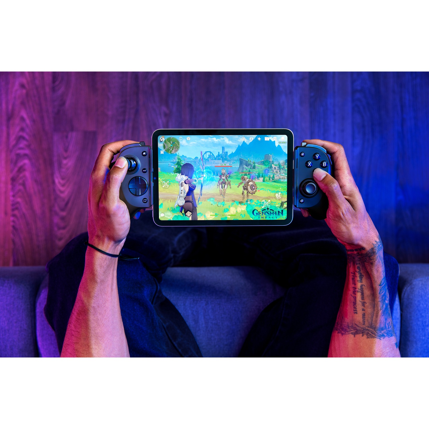 Hands holding a tablet with game controllers attached. The screen shows a game with characters. Tattoo on the arm. Seated on a sofa.