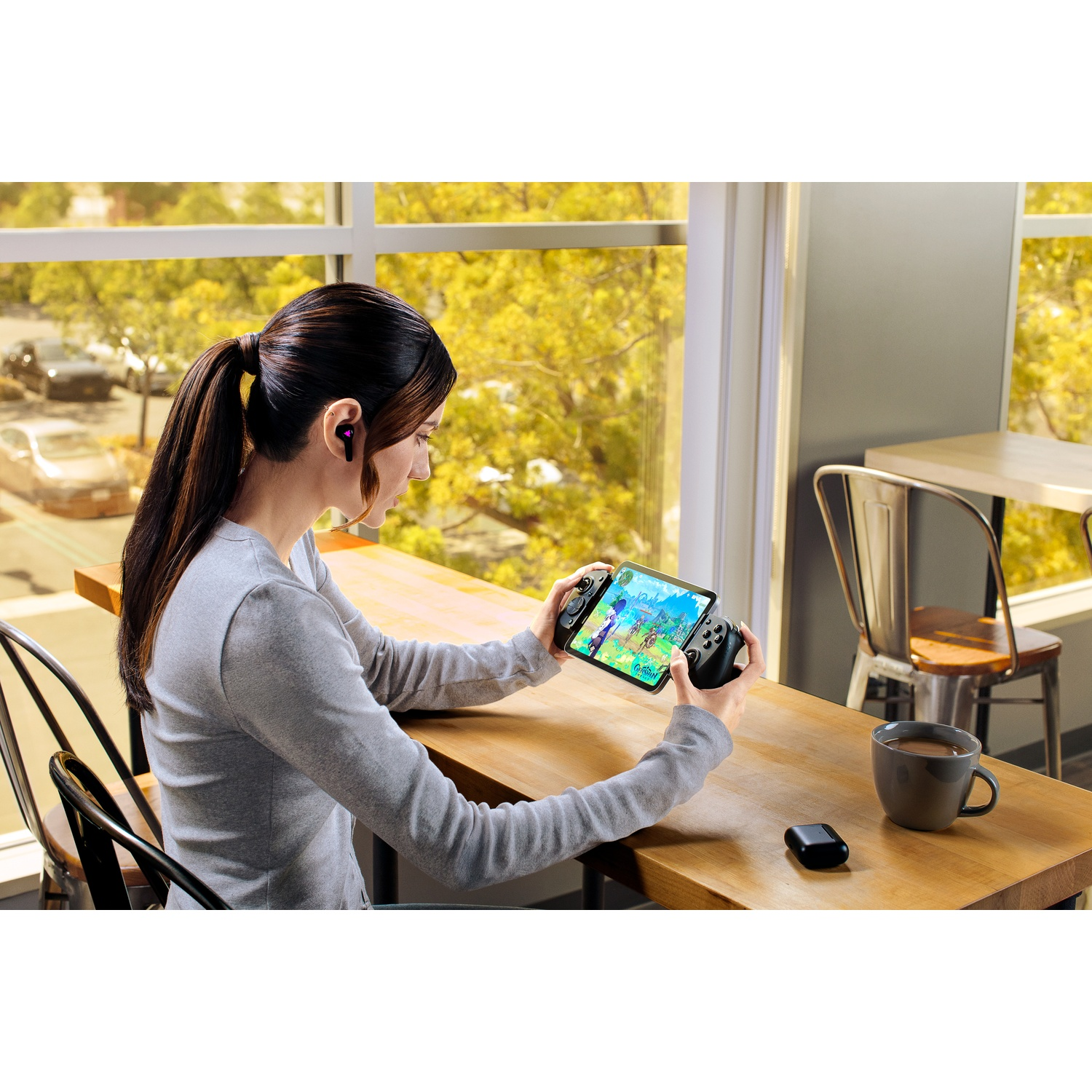Woman with earbuds plays a game on a tablet attached to controllers. She sits at a table near a window with a coffee cup.