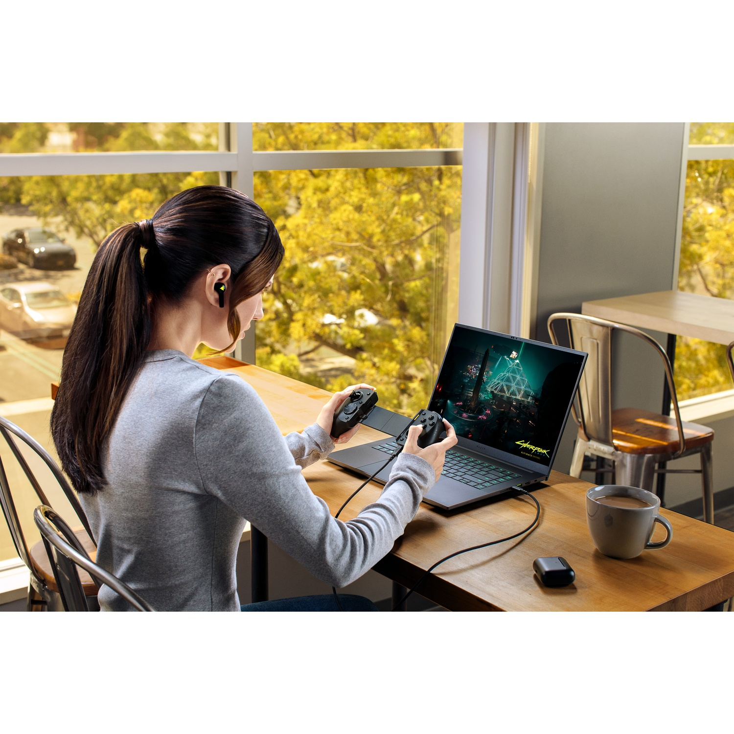 Woman gaming on a laptop with controllers. She wears earbuds and sits at a wooden table. Outside, trees and cars are visible.