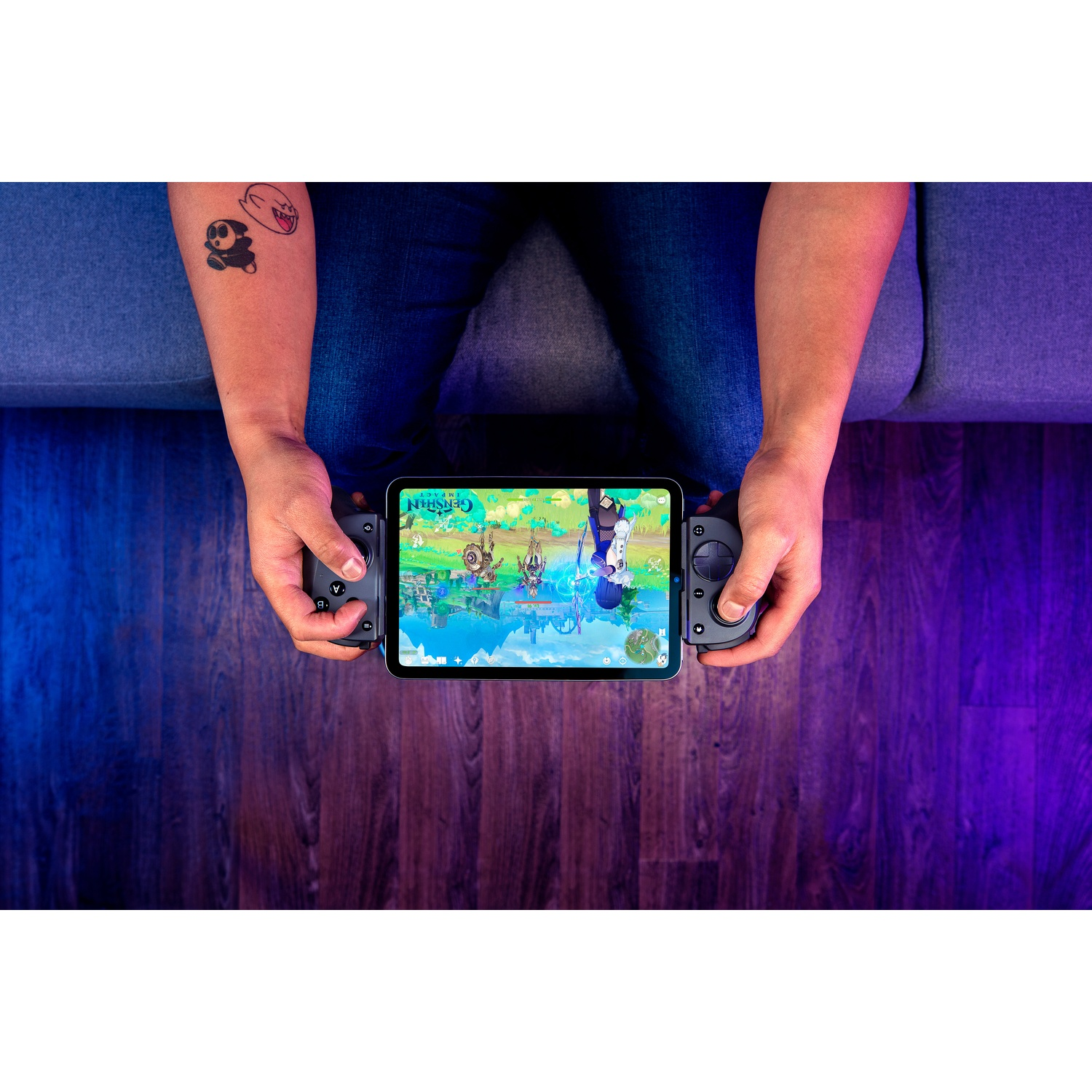 A person is playing a game on a tablet with a controller. The tablet shows a game with characters. There is a tattoo on the arm.