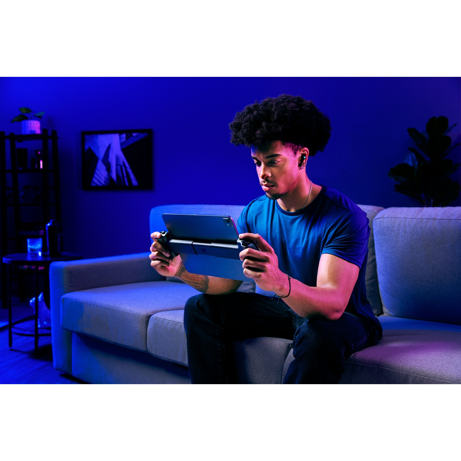 Een man met oortelefoons speelt op een tablet met een controller, zittend op een grijze bank. De kamer is zwak blauw verlicht.