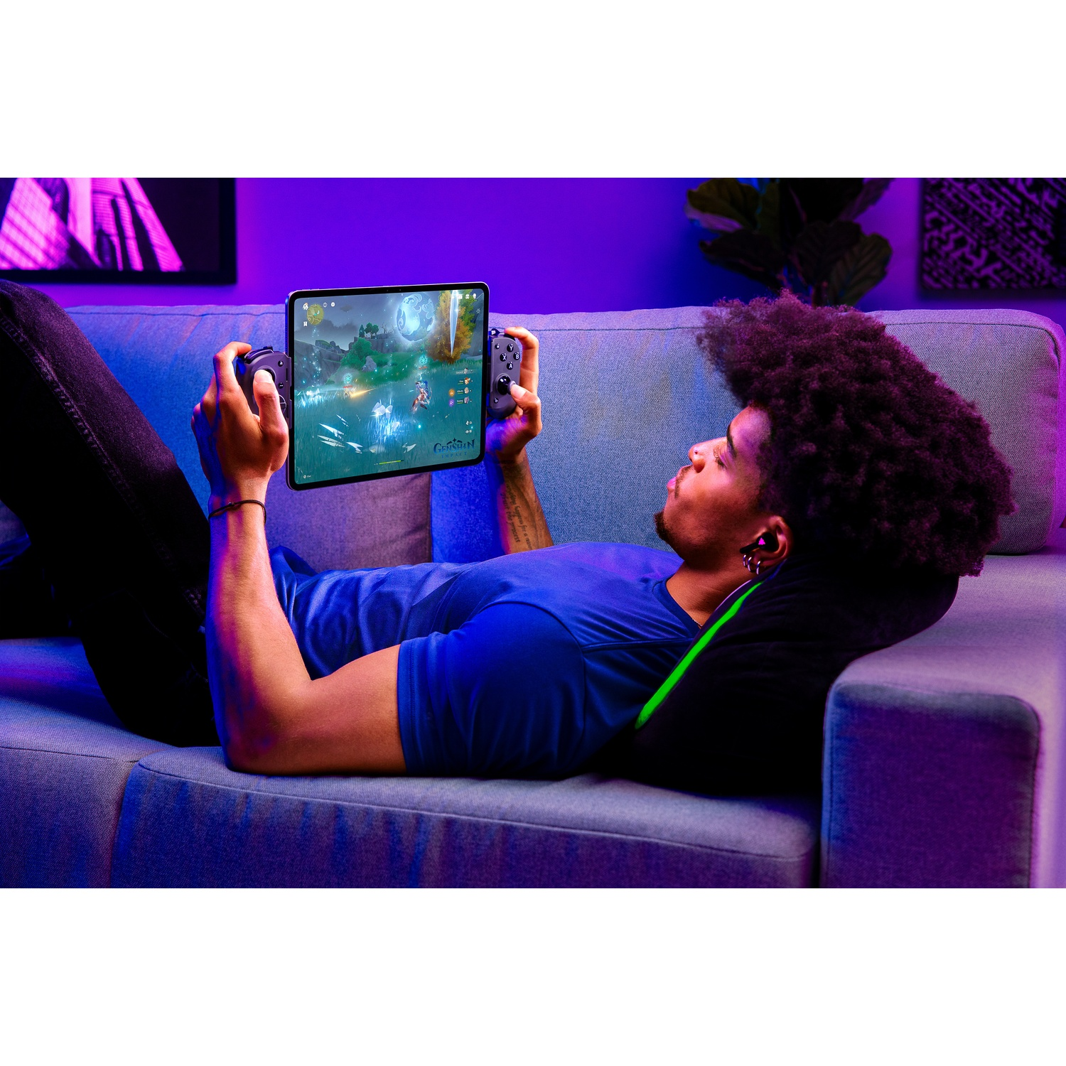 Een man ligt op een bank en speelt een spel op een tablet met controllers. De kamer is zwak verlicht, met een paarse tint.