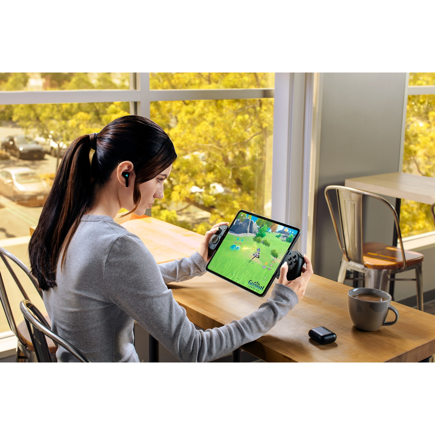 Vrouw speelt een game op een tablet met controller, draagt oortjes, aan een tafel bij een raam. Koffiekopje en etui staan op tafel.