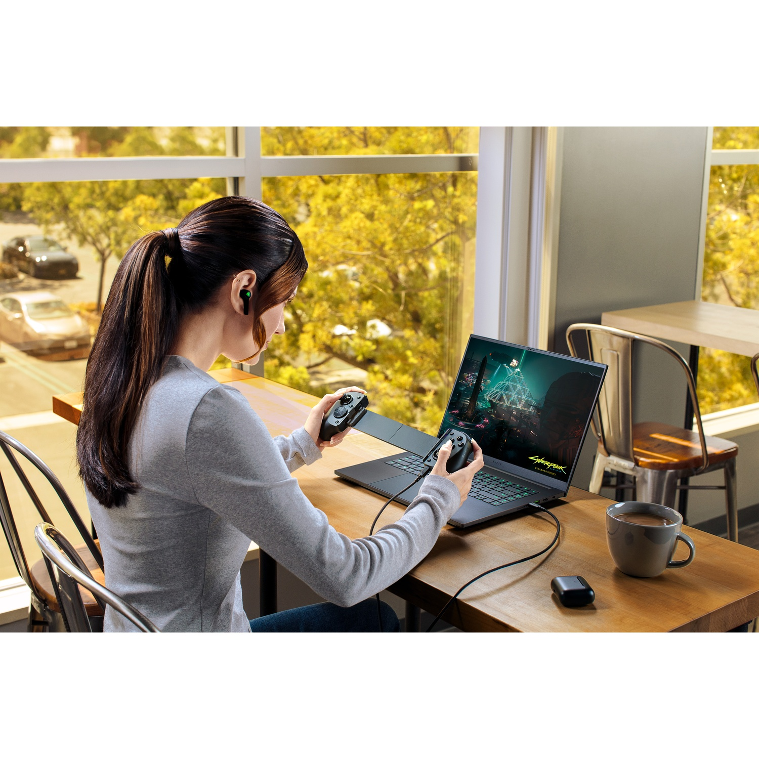 Vrouw speelt videogame op laptop, houdt controllers vast, draagt oordopjes. Koffiekopje en oplader op tafel. Auto's zichtbaar buiten.