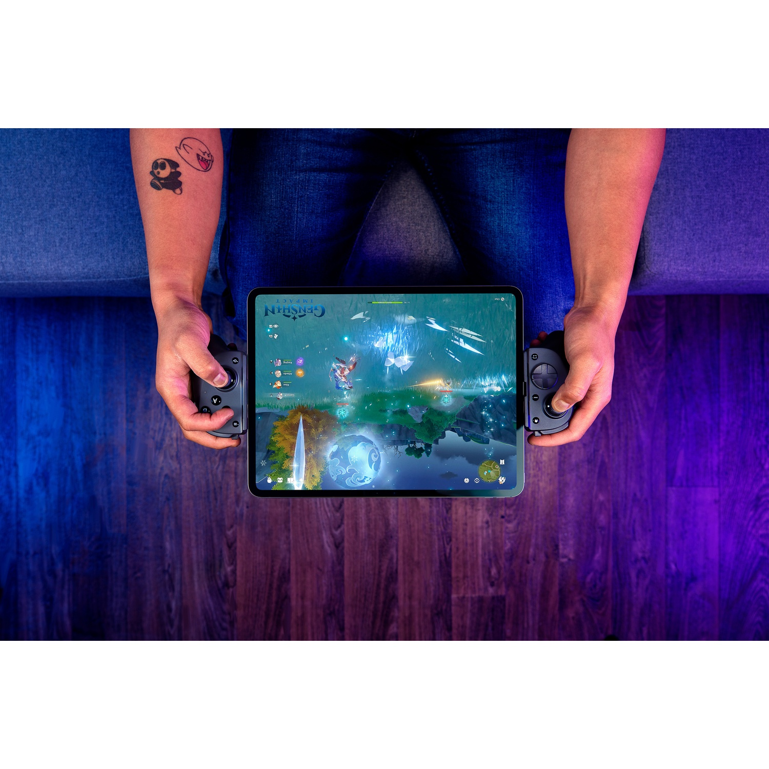 Een persoon speelt een spel op een tablet met aangesloten controllers. Ze zitten op een bank. Een tatoeage is zichtbaar op hun arm.