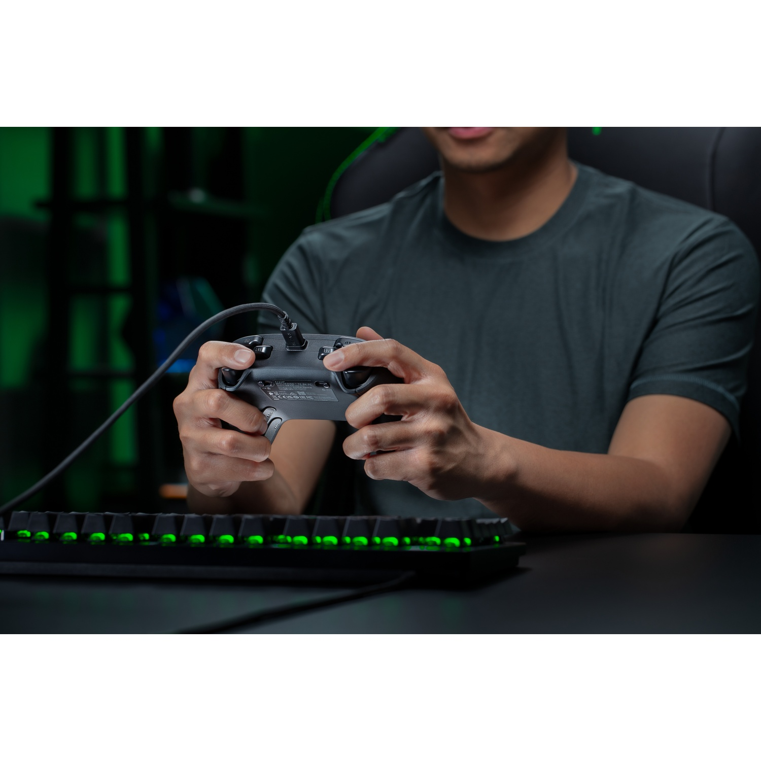 A person is holding a gray controller connected to a black keyboard with green lights. The background is dark.