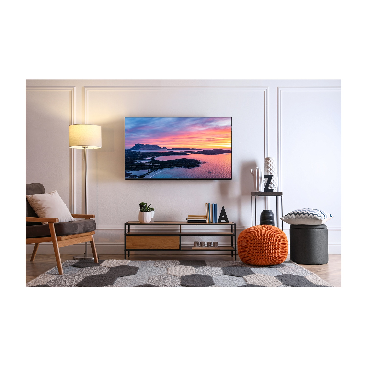 Soggiorno con TV. Sedia, tappeto, pouf arancione e tavolino. La TV mostra un paesaggio al tramonto. Parete bianca con modanature.