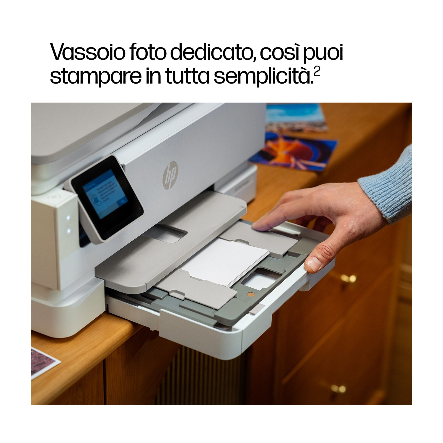 Una mano inserisce un vassoio di carta in una stampante HP. La stampante è su una superficie di legno, con una foto visibile nelle vicinanze.