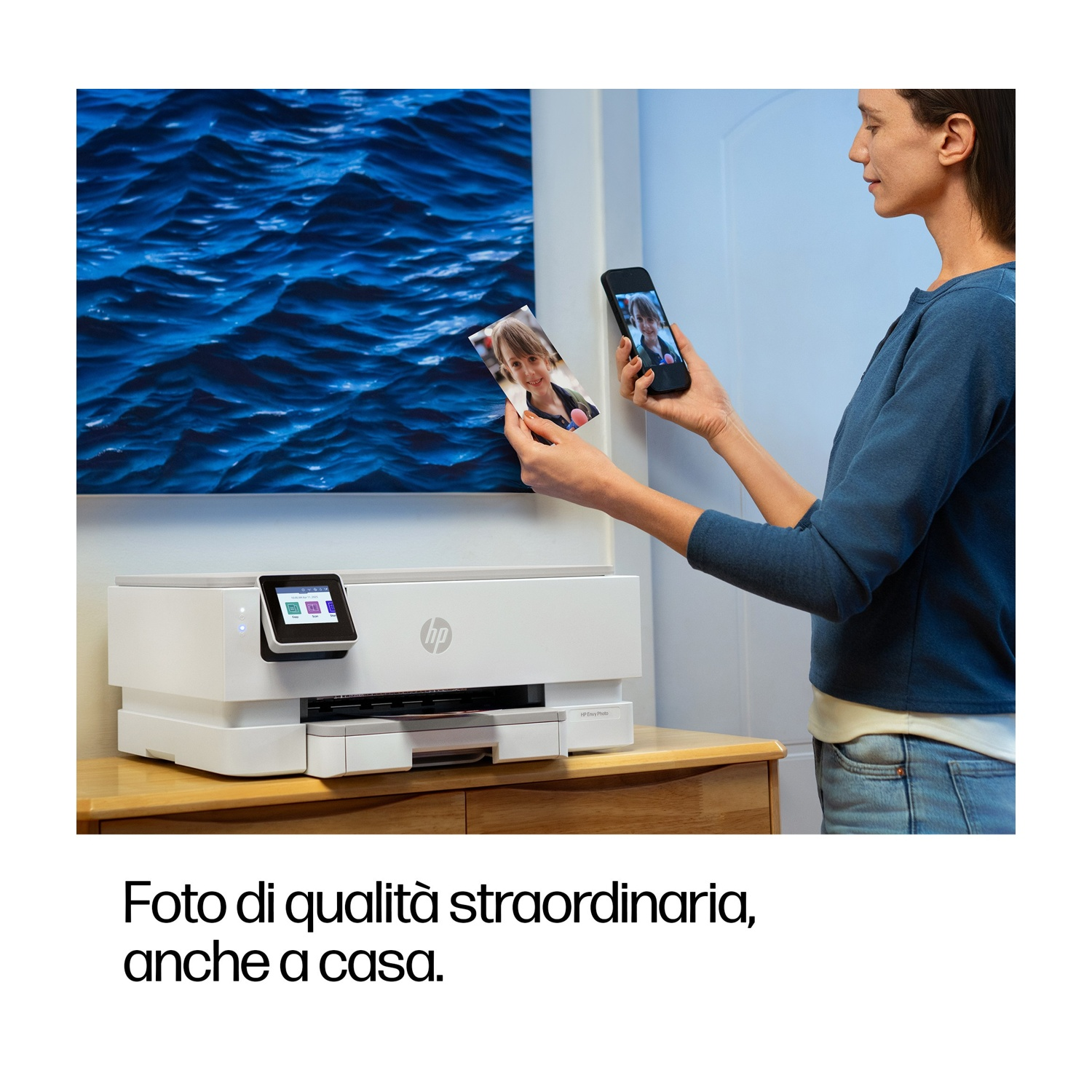 Donna confronta foto stampata con immagine del telefono. Stampante bianca su tavolo di legno, arte oceanica blu dietro.