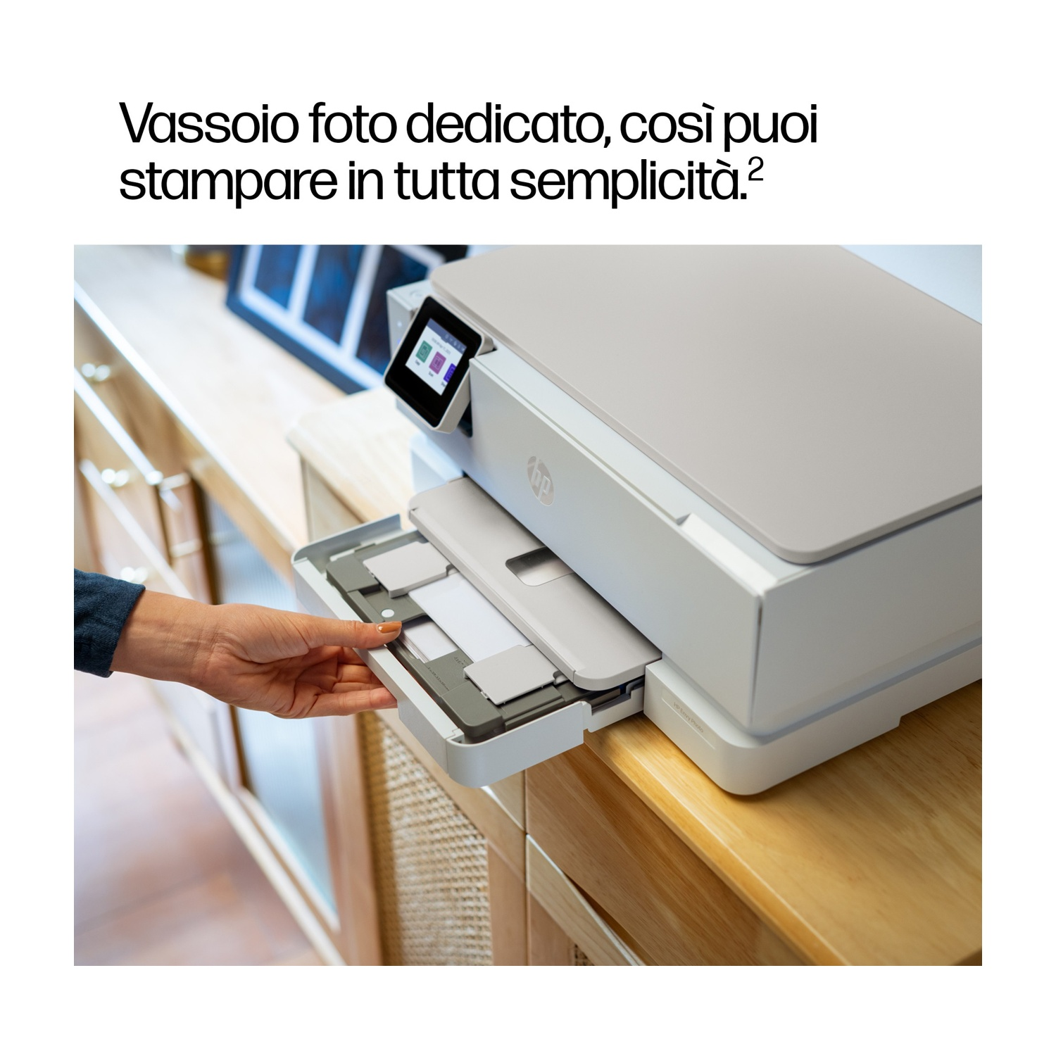 Una mano apre un vassoio della stampante. La stampante è bianca e grigia. È su un mobile di legno, testo in alto.