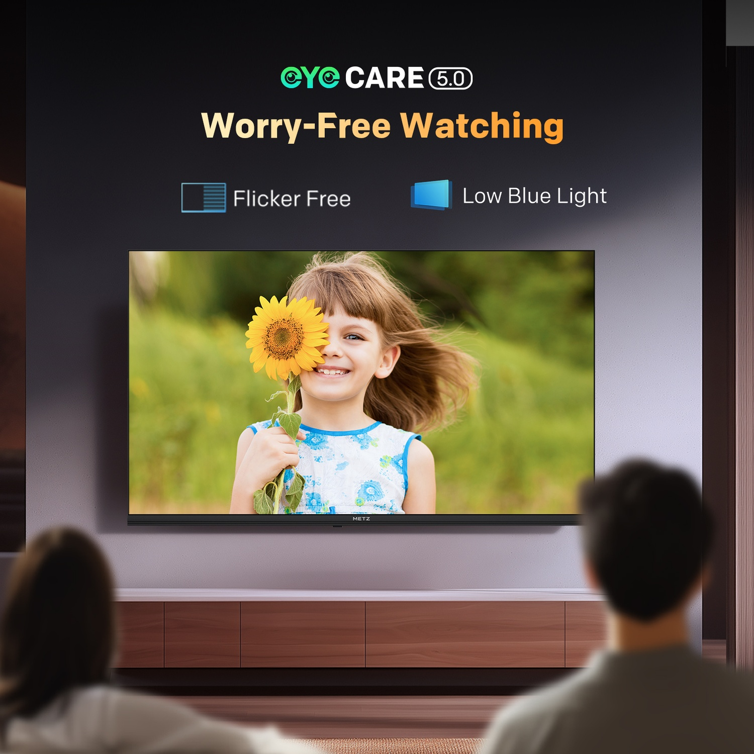 Un televisore mostra una bambina con un girasole. Sopra, il testo dice 'Visione senza preoccupazioni'. Due persone in primo piano.