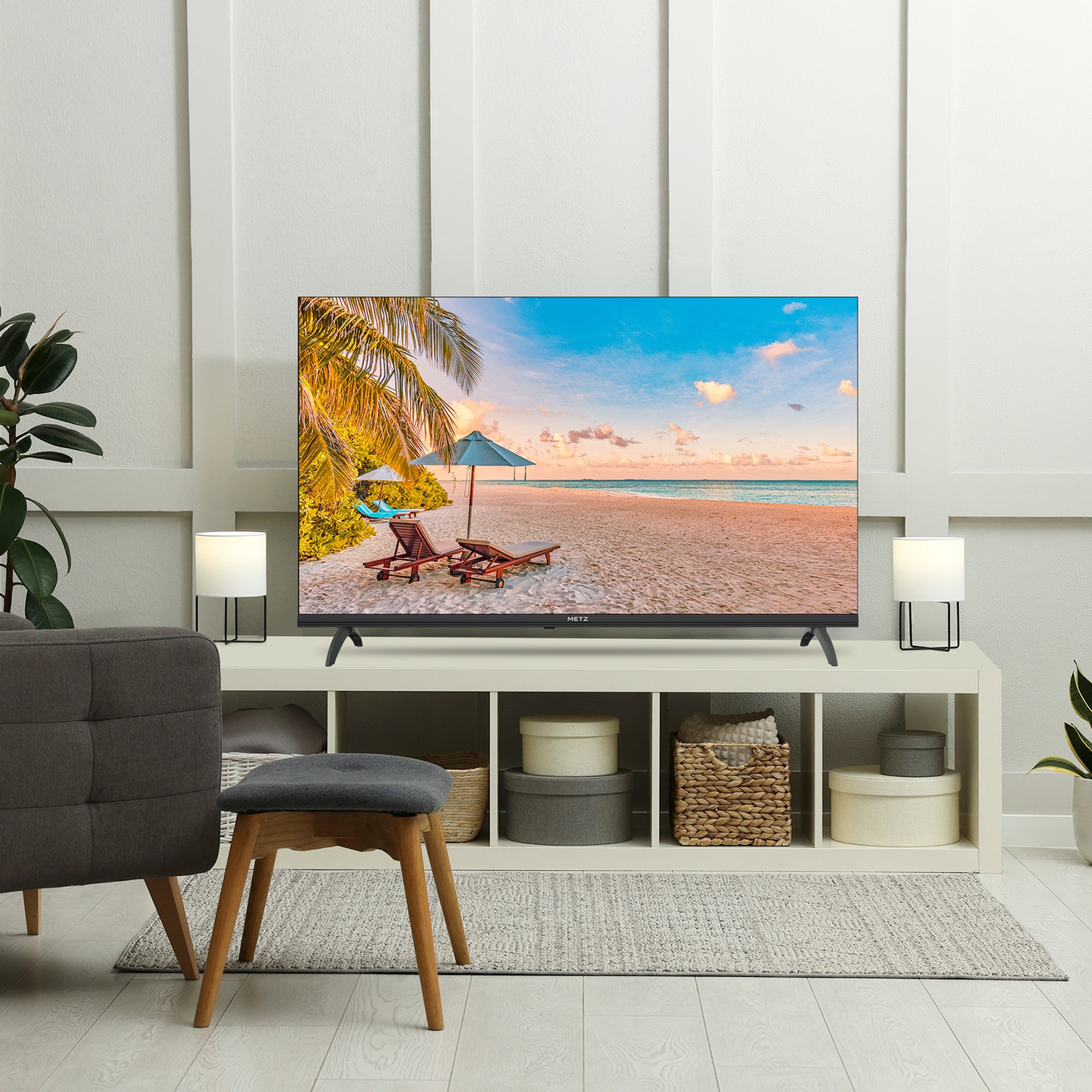 La TV mostra una scena di spiaggia. La stanza ha una sedia grigia, uno sgabello e un contenitore bianco. Lampade sullo scaffale. Piante e scatole.