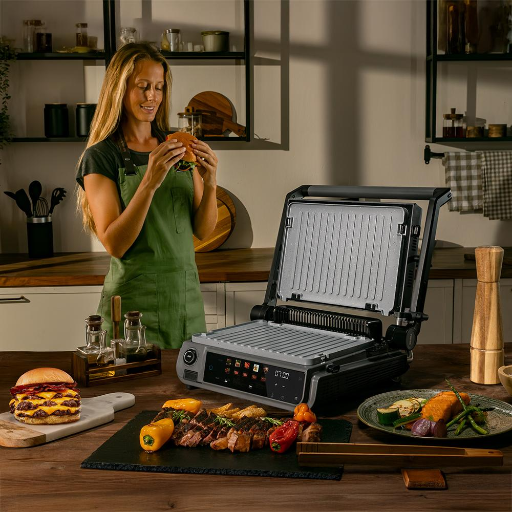 Mujer con delantal verde sosteniendo una hamburguesa. Una parrilla, comida y artículos de cocina están sobre una mesa de madera. Estantes y cocina al fondo.