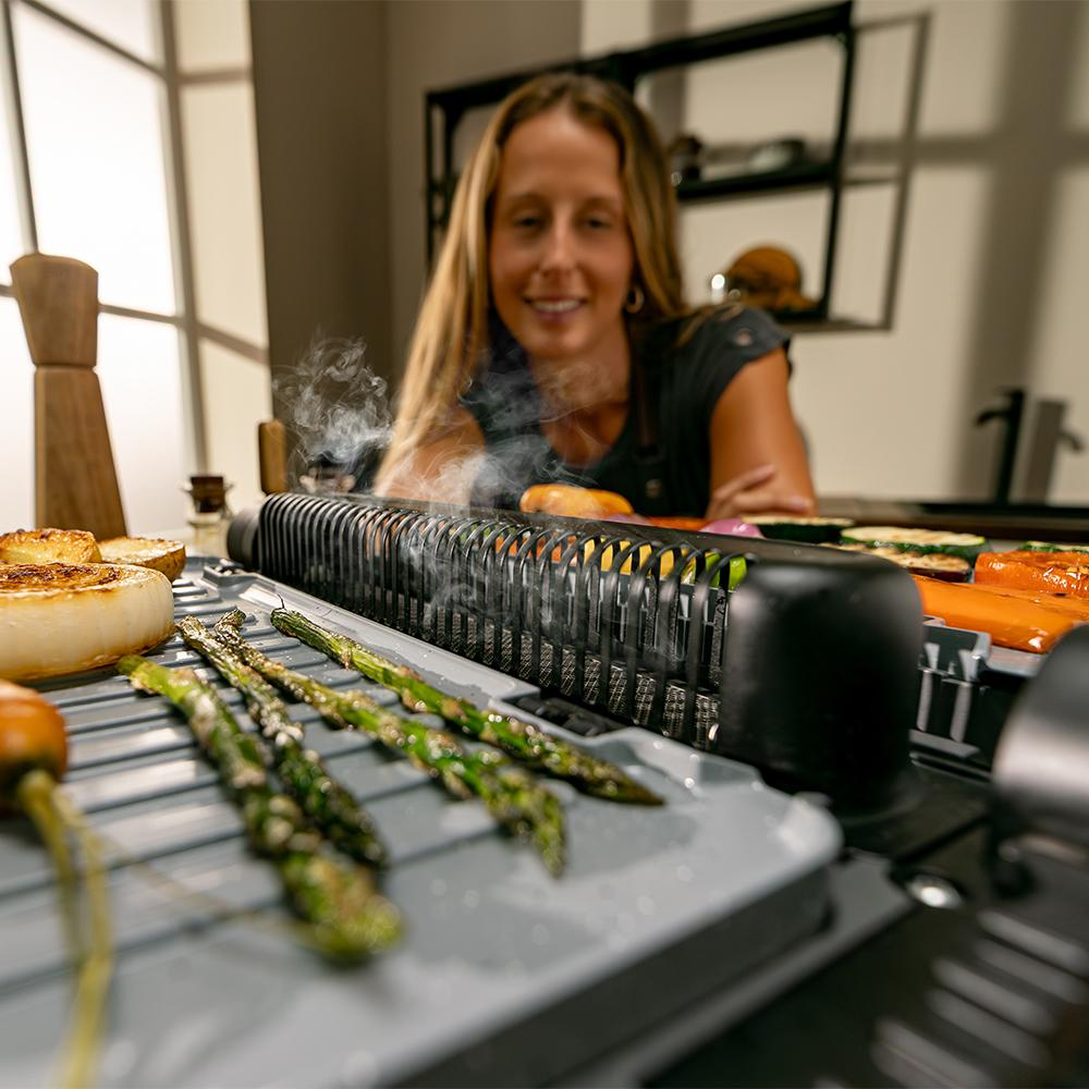 Una mujer sonríe sobre una parrilla con verduras. Se ven espárragos, cebollas y zanahorias. El vapor se eleva de la parrilla.