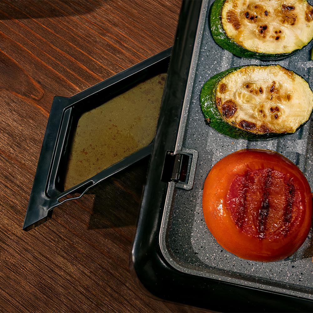 Verduras a la parrilla en una parrilla. Se ven calabacín y tomate. Una bandeja negra está a la izquierda. Mesa de madera.