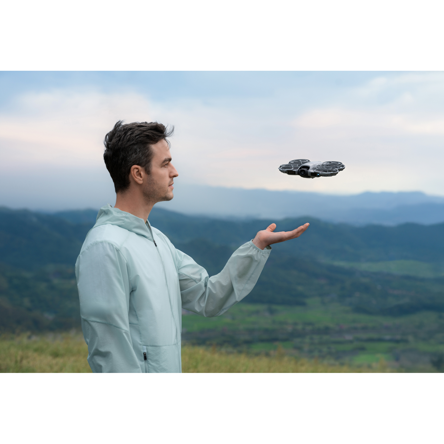 Een man in een lichte jas houdt zijn hand uit naar een drone in de lucht. Bergen en lucht op de achtergrond.