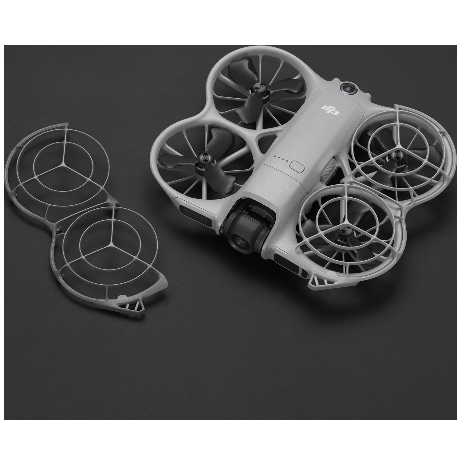 Een grijze drone met beschermende kooien en zwarte propellers ligt op een zwart oppervlak. Een losse kooi is in de buurt. Het DJI-logo is zichtbaar.