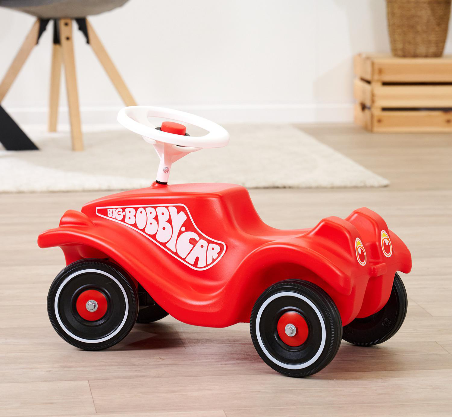 Ein rotes Big Bobby Car-Spielzeug steht auf einem Holzboden. Es hat ein weißes Lenkrad. Im Hintergrund befinden sich Möbel und ein Teppich.