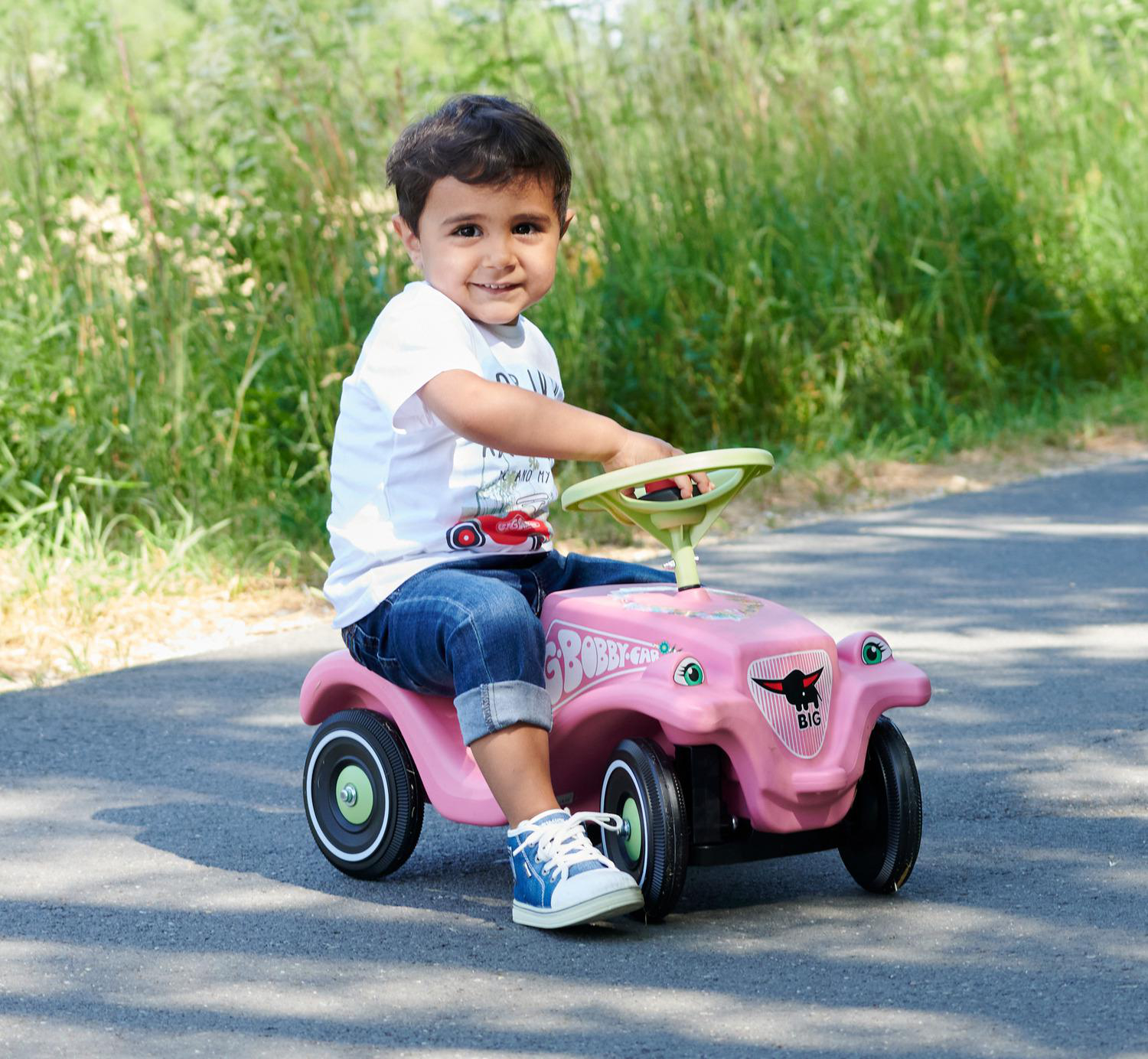 Ein Junge sitzt lächelnd auf einem rosa Spielzeugauto. Er trägt Jeans, ein weißes Hemd und blaue Turnschuhe. Grünes Gras im Hintergrund.