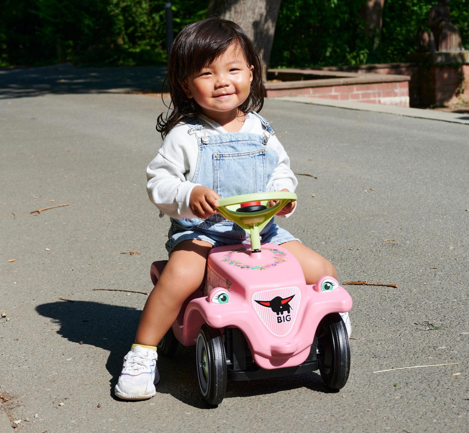 Ein lächelndes Kind auf einem rosa Spielzeugauto. Das Kind trägt eine Jeanslatzhose und sitzt auf dem Auto auf einer Asphaltstraße. Bäume sind im Hintergrund.
