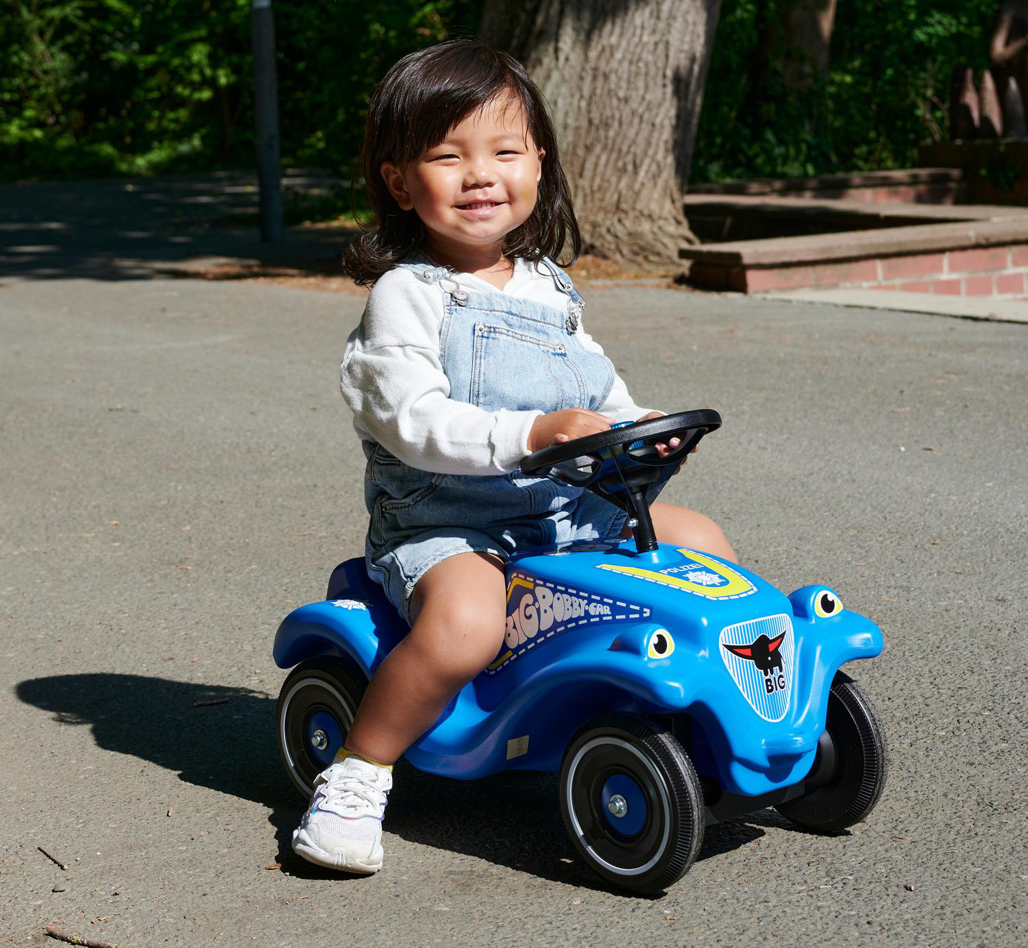 Ein lächelndes Kind sitzt auf einem blauen Spielzeugauto. Der Hintergrund umfasst Bäume und Pflaster.