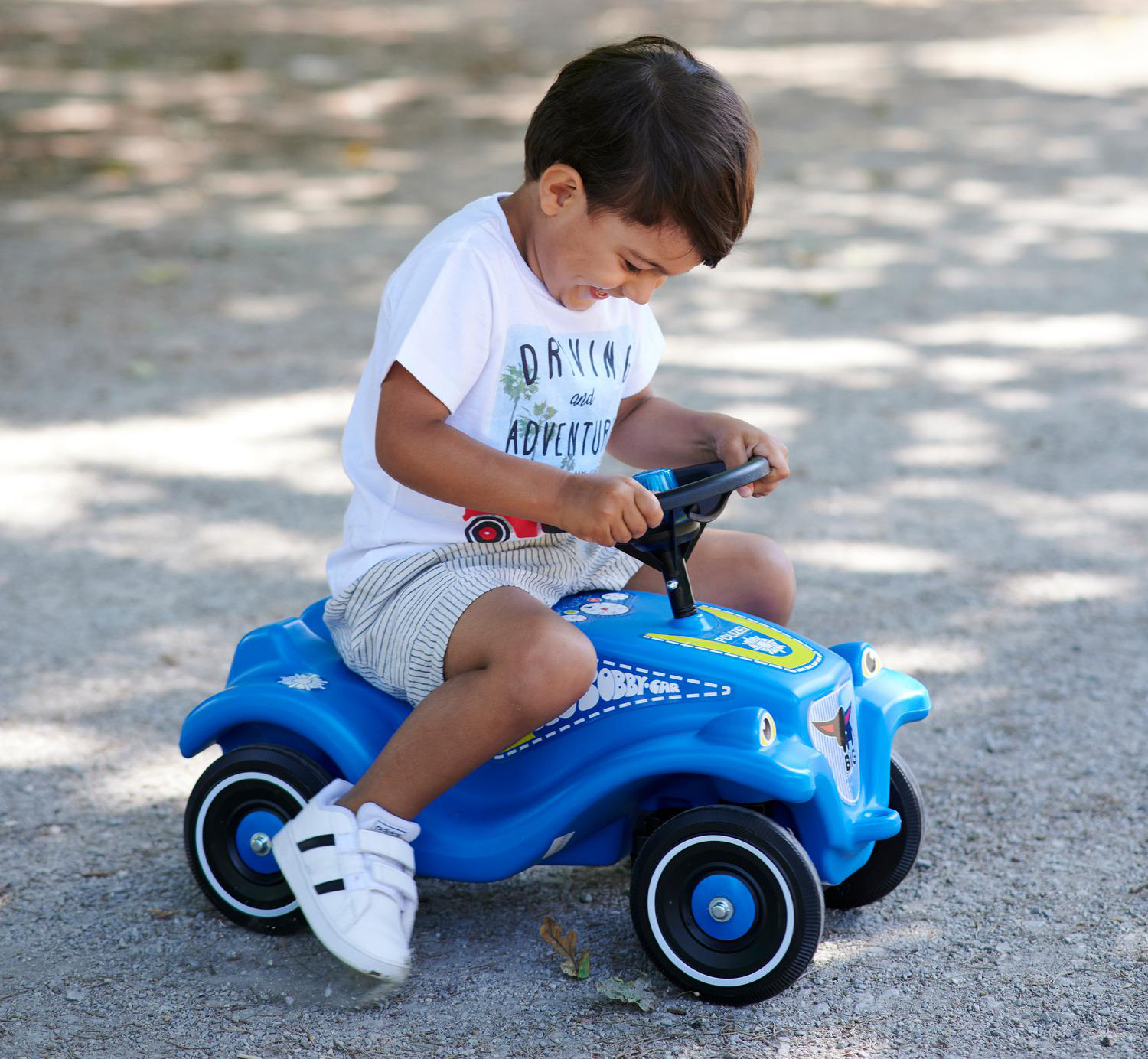 Ein kleiner Junge sitzt auf einem blauen Spielzeugauto. Er trägt ein weißes Hemd und Shorts. Das Auto steht auf einem Schotterweg.