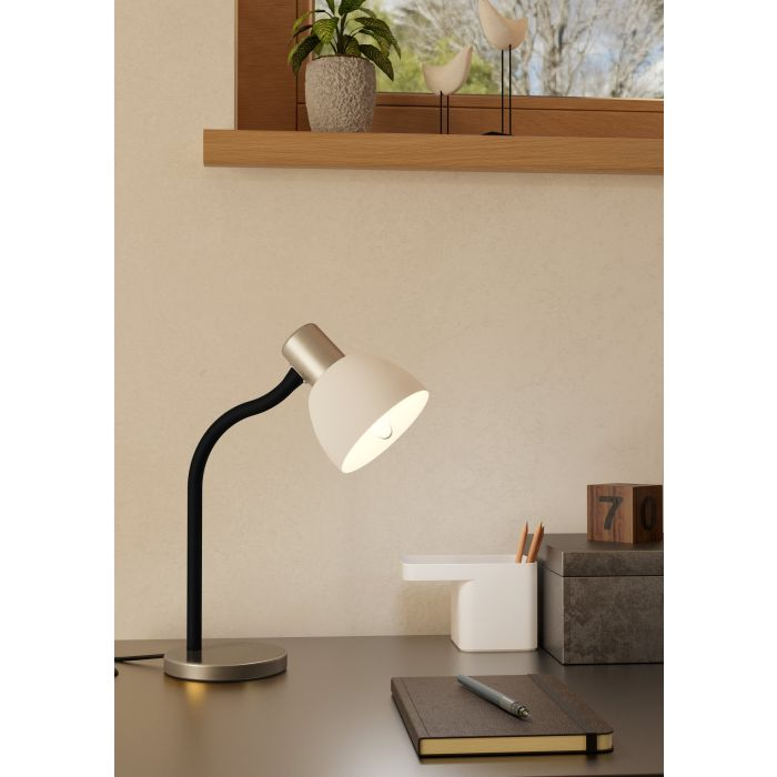 Eine Schreibtischlampe mit weißem Lampenschirm leuchtet. Auf dem Schreibtisch liegen ein Notizbuch und ein Stift. Im Hintergrund befindet sich ein Fenster mit Pflanzen und Dekoration.
