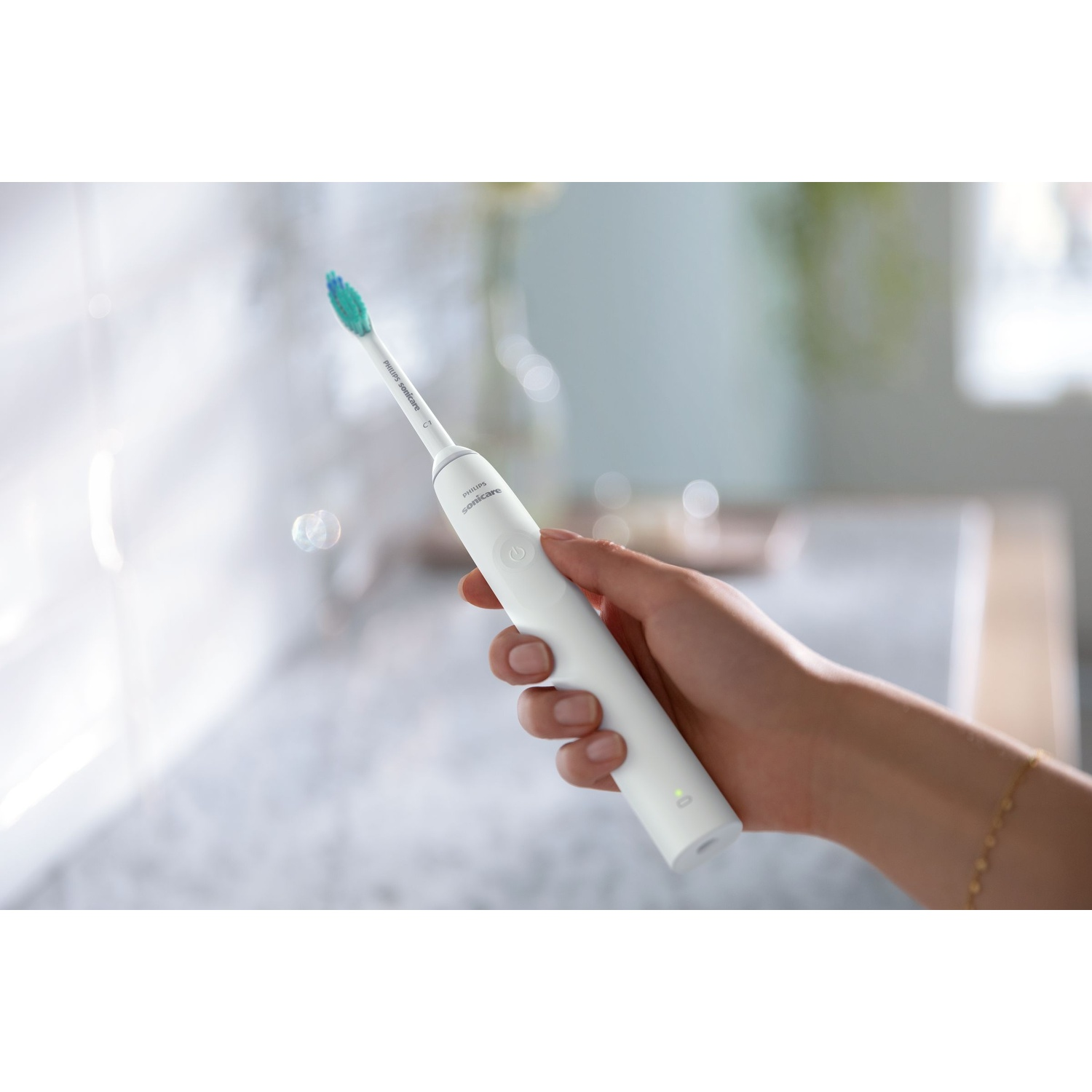 Una mano tiene in mano uno spazzolino elettrico bianco con una testina verde e blu. Lo sfondo è un bagno.
