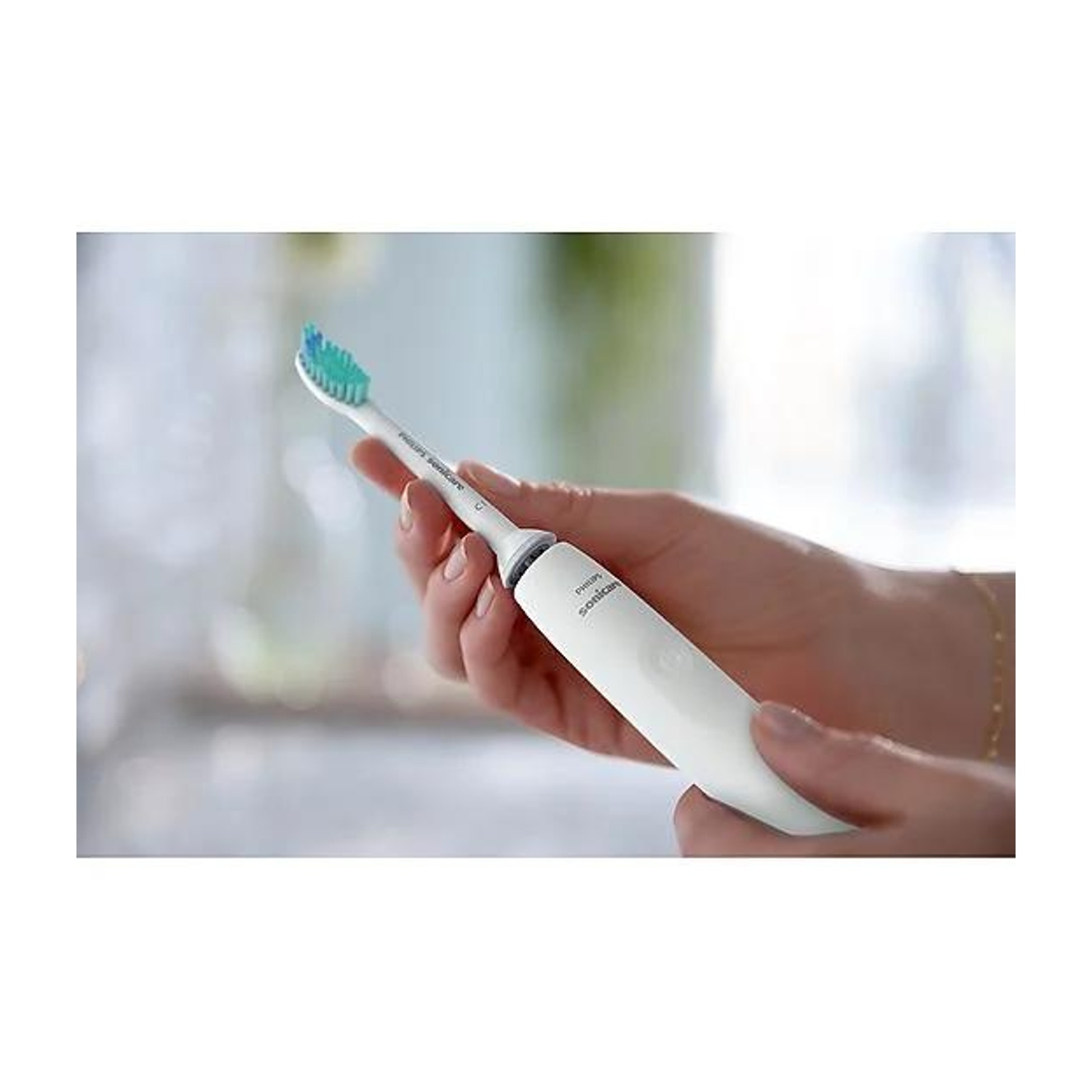 Una persona tiene in mano uno spazzolino elettrico bianco con setole verdi e blu. Lo sfondo è sfocato, mostrando una finestra.