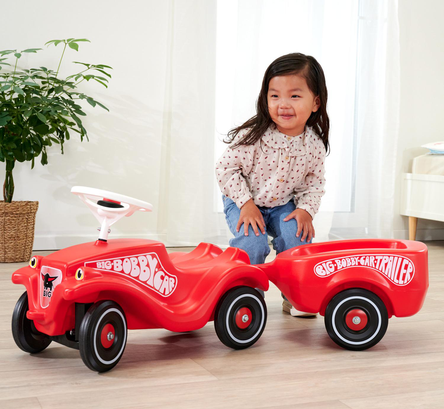 Ein lächelndes Kind steht in der Nähe eines roten Spielzeugautos mit Anhänger. Links steht eine Pflanze in einem Weidenkorb. Helles Zimmer.