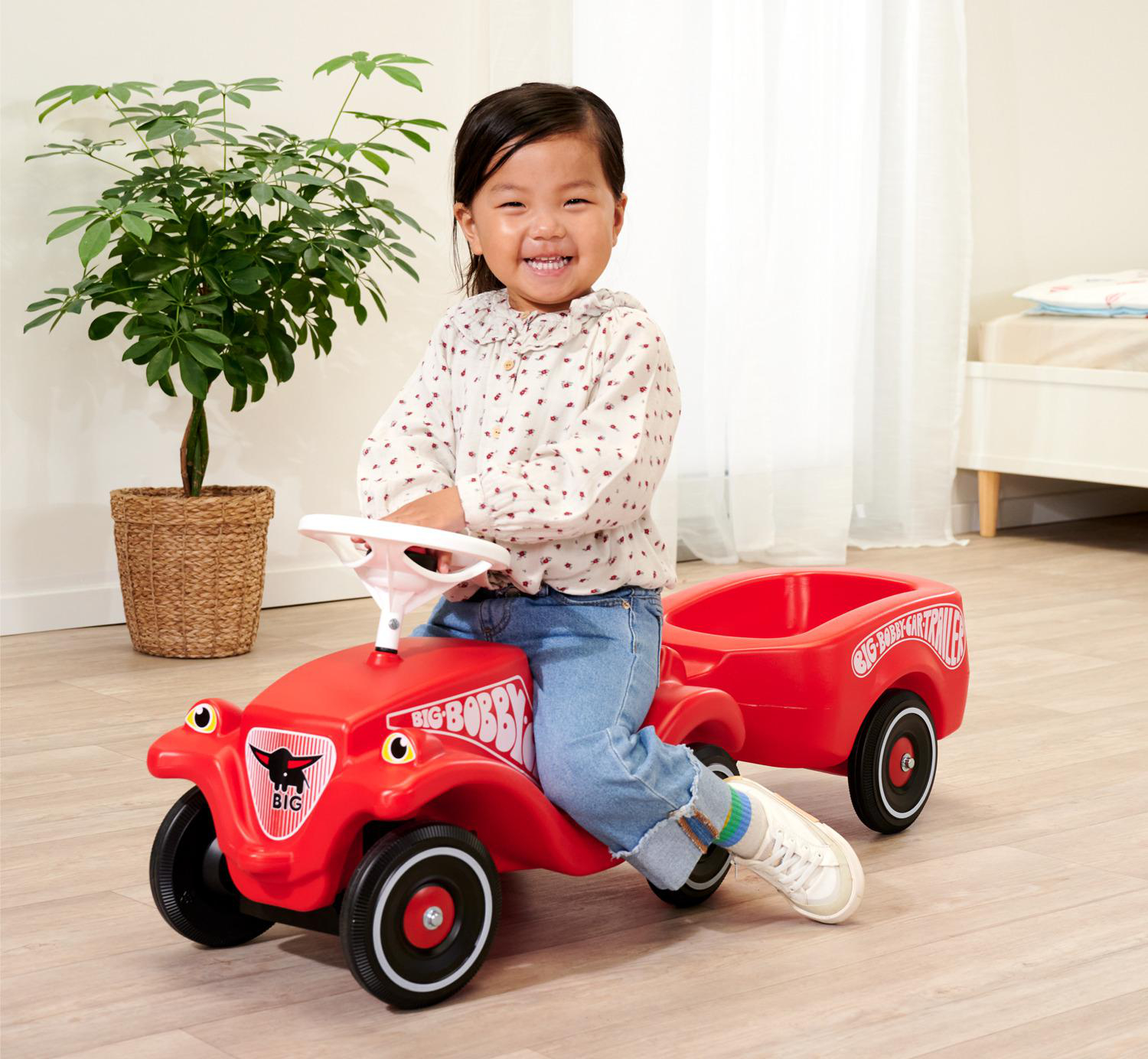Ein lächelndes Kind sitzt auf einem roten Spielzeugauto mit Anhänger. Links ist eine Pflanze im Korb, im Hintergrund ein Bett.