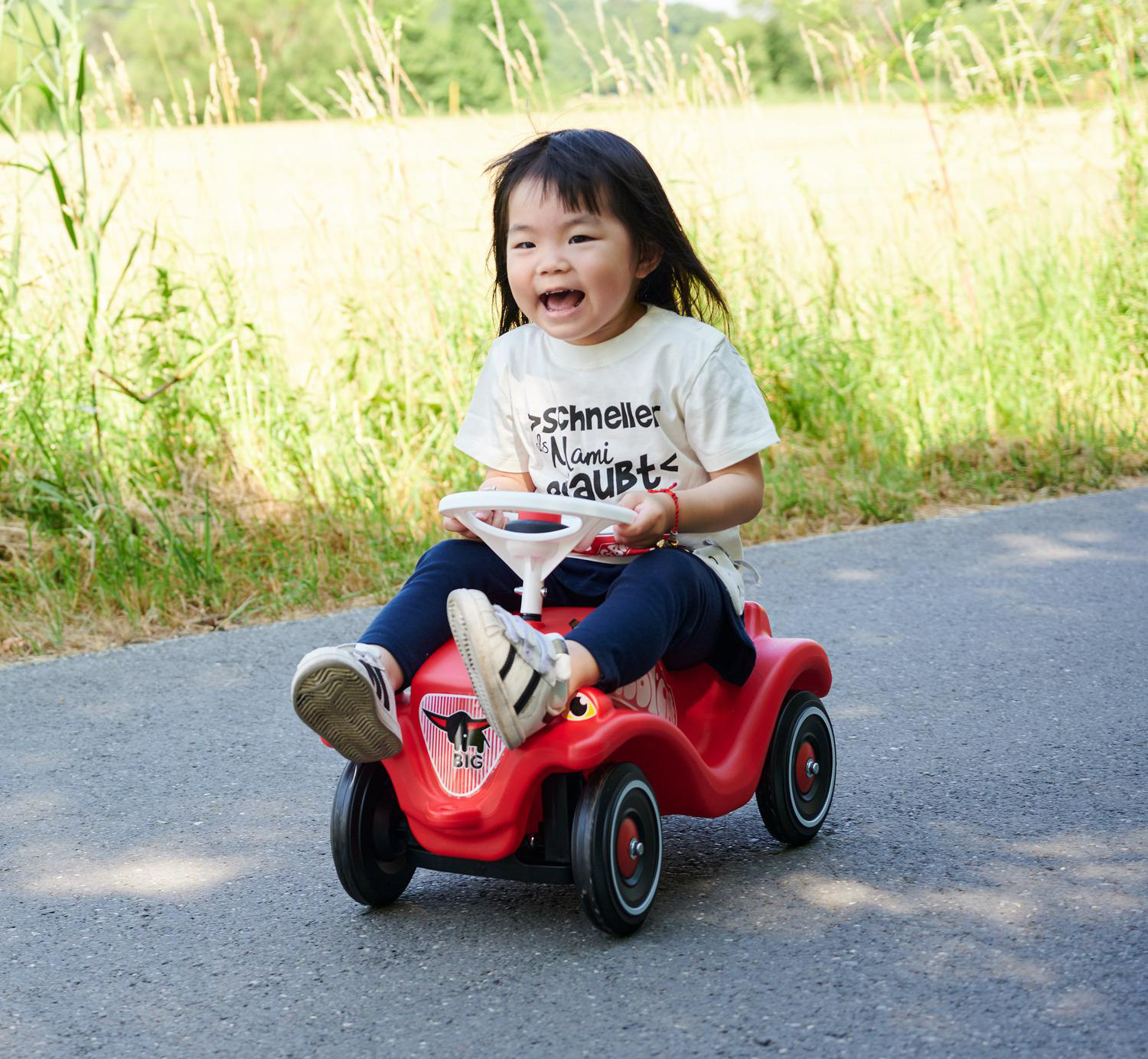 Ein Kind fährt fröhlich mit einem roten Spielzeugauto auf einer Straße. Das Kind trägt ein weißes Hemd und dunkle Hosen, im Hintergrund ein grünes Feld.
