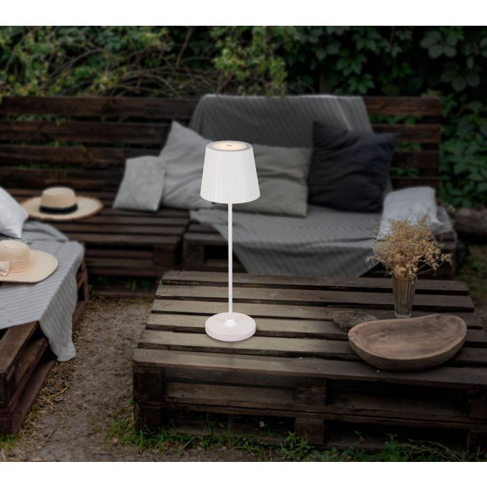Eine weiße Lampe steht auf einem Holztisch. Im Hintergrund sind ein Sofa und Hüte zu sehen. Die Szene spielt sich im Freien ab.