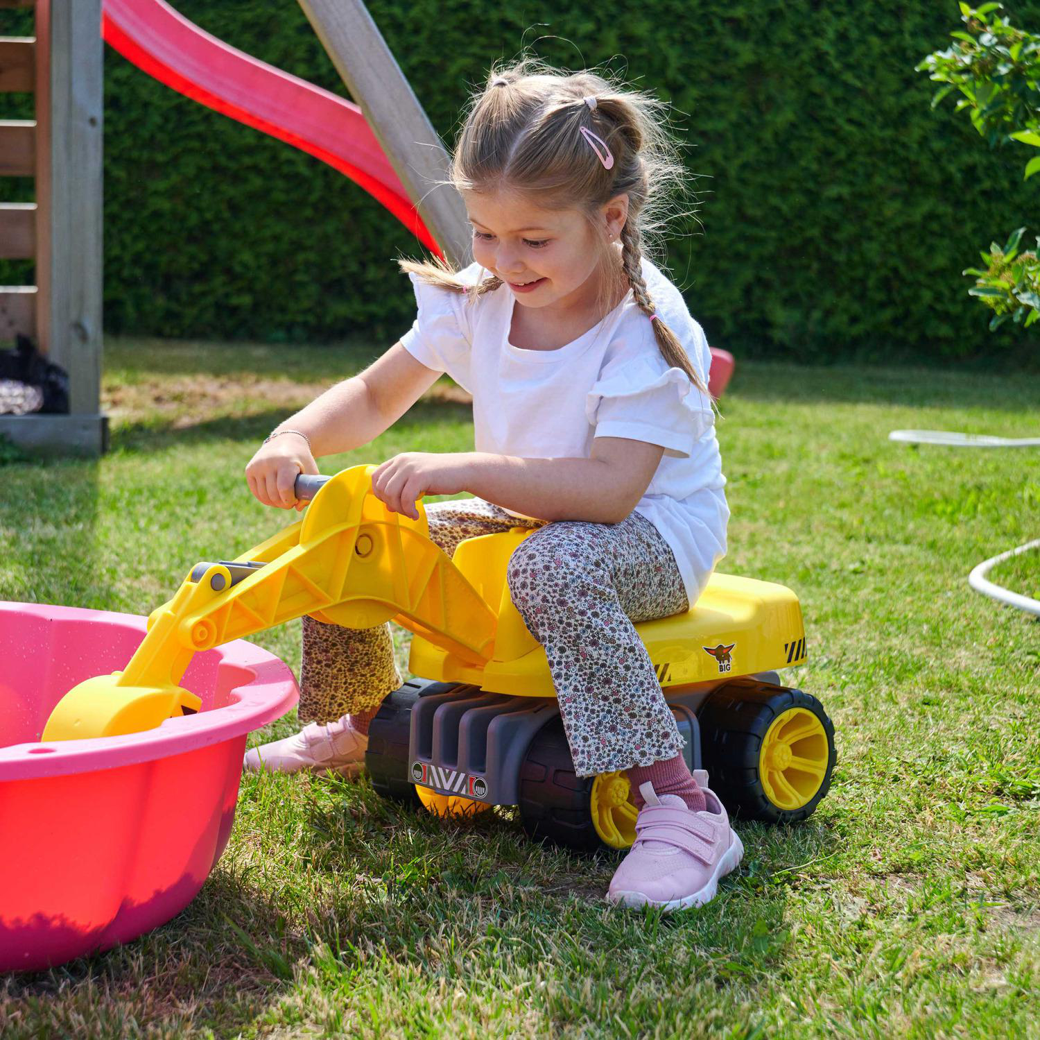 Ein Mädchen auf einem gelben Spielzeugbagger. Sie lächelt, in der Nähe einer roten Wanne und einer Rutsche auf einer Rasenfläche. Grüne Büsche im Hintergrund.