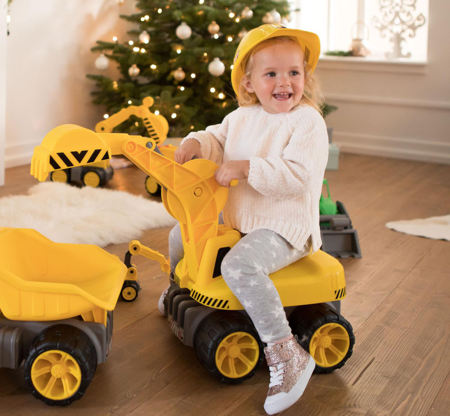 Ein junges Mädchen mit gelbem Bauhelm lächelt, während es auf einem gelben Spielzeugbagger spielt. Im Hintergrund steht ein Weihnachtsbaum.