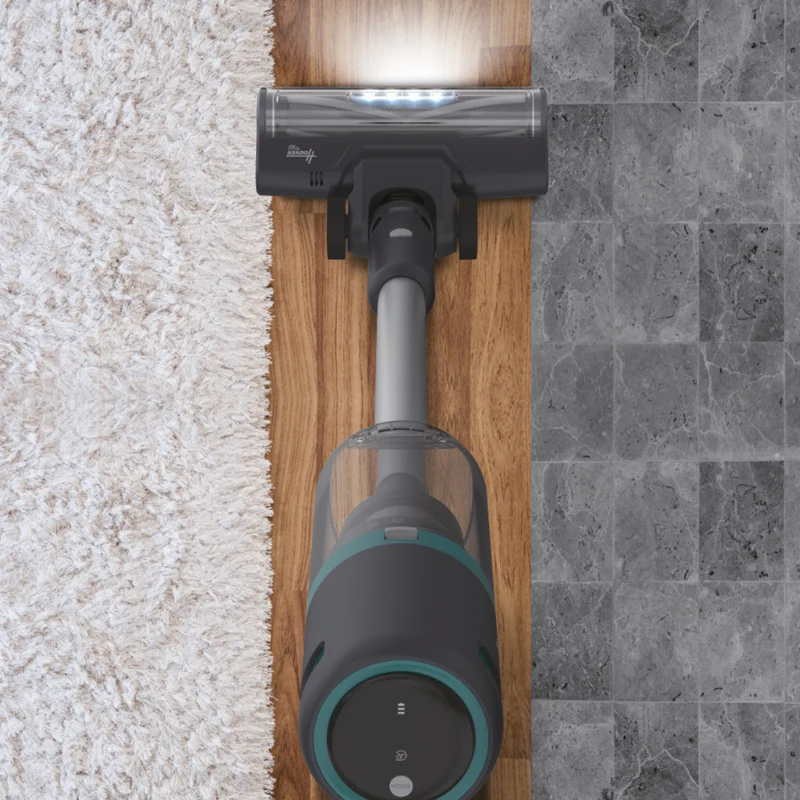 Un aspirapolvere senza fili su pavimento in legno e piastrelle. L'aspirapolvere è grigio scuro con dettagli turchese. Tappeto bianco a sinistra.
