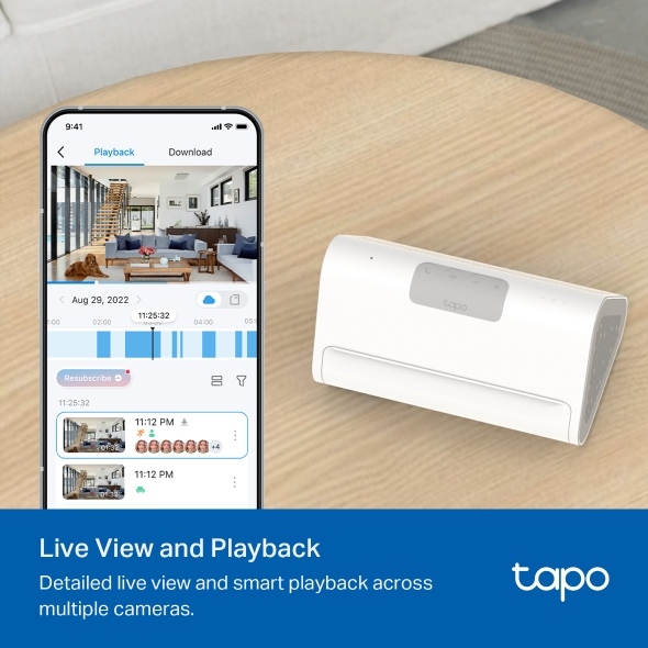 Ein Smartphone zeigt ein Video vom Innenraum eines Hauses. Auf einer Holzoberfläche befindet sich ein weißer Lautsprecher. Das Tapo-Logo ist unten rechts.