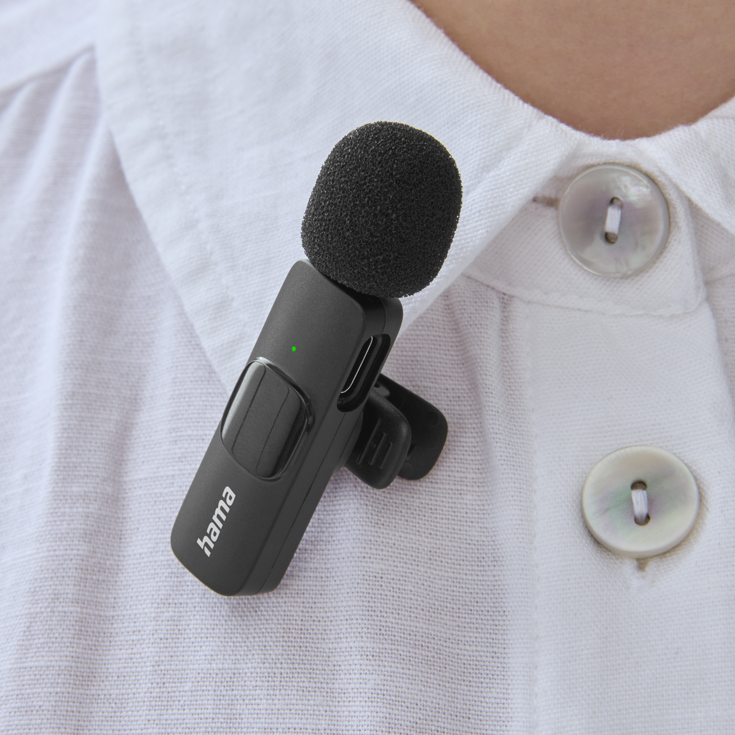 A black microphone is clipped to a white shirt. The microphone is small with a foam head. A green light is visible on it.