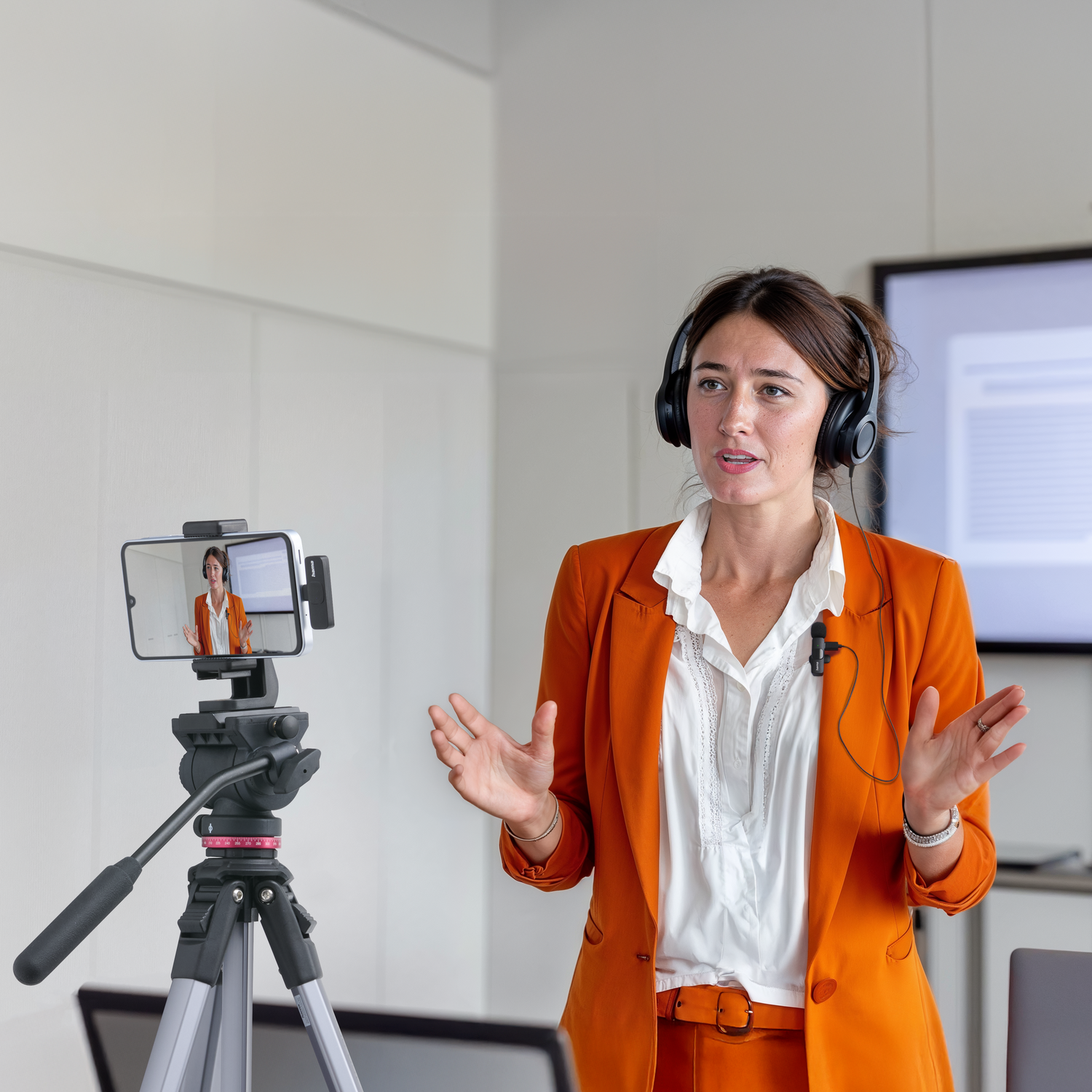 Woman in orange suit and headphones speaking. Phone on tripod records her. White shirt and background. 