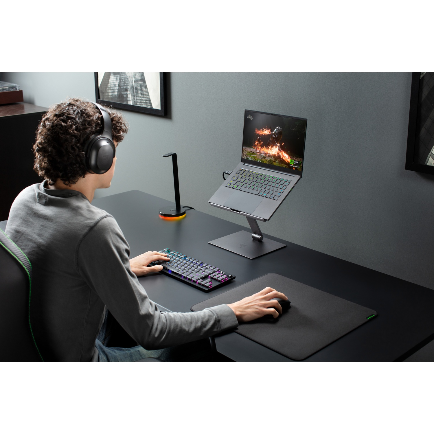 A person with headphones is using a laptop on a desk. Keyboard and mouse are in front. A lamp is on the desk.