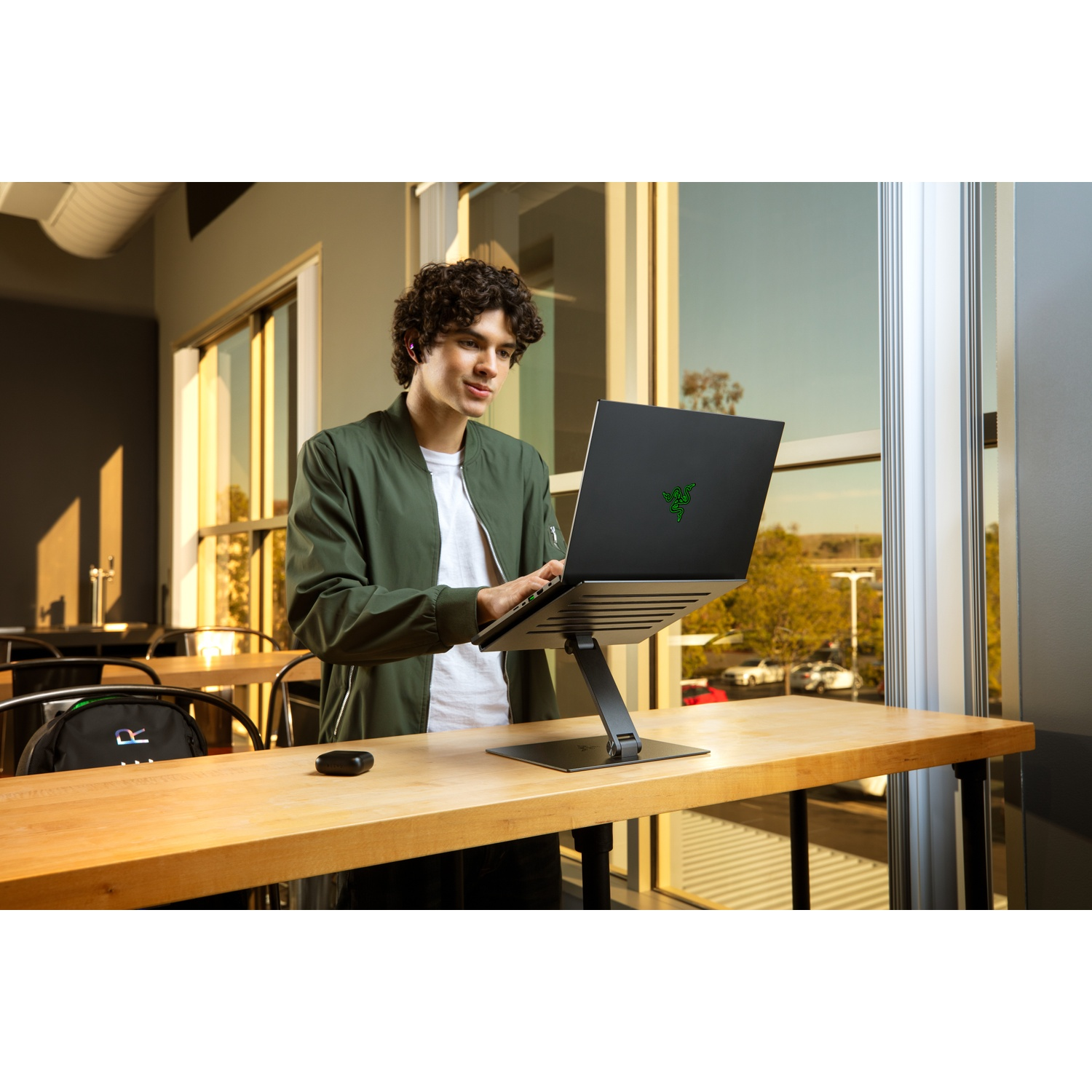 A man stands using a laptop on a stand. He wears a green jacket. The laptop has a green logo.