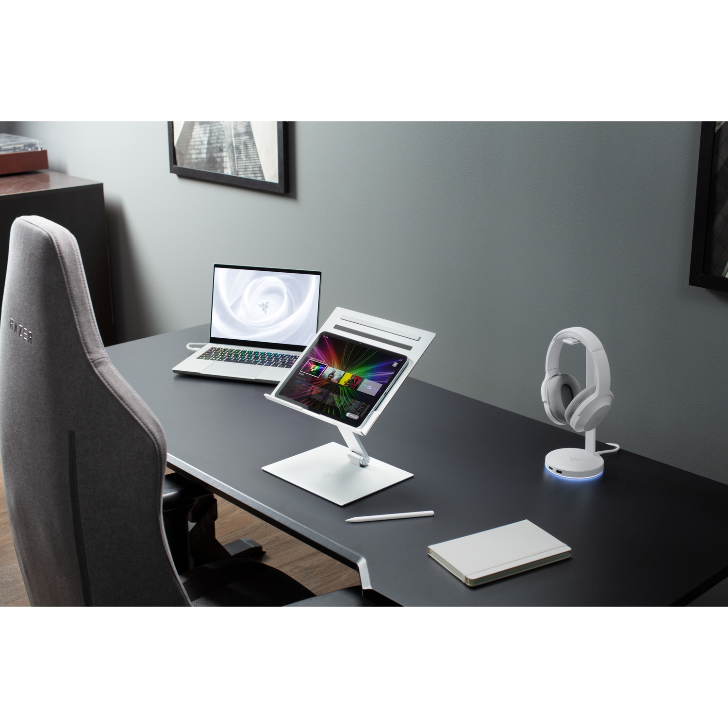 A desk with a laptop, tablet, headphones, and notebook. The laptop and tablet are on stands. A pen lies next to the notebook.