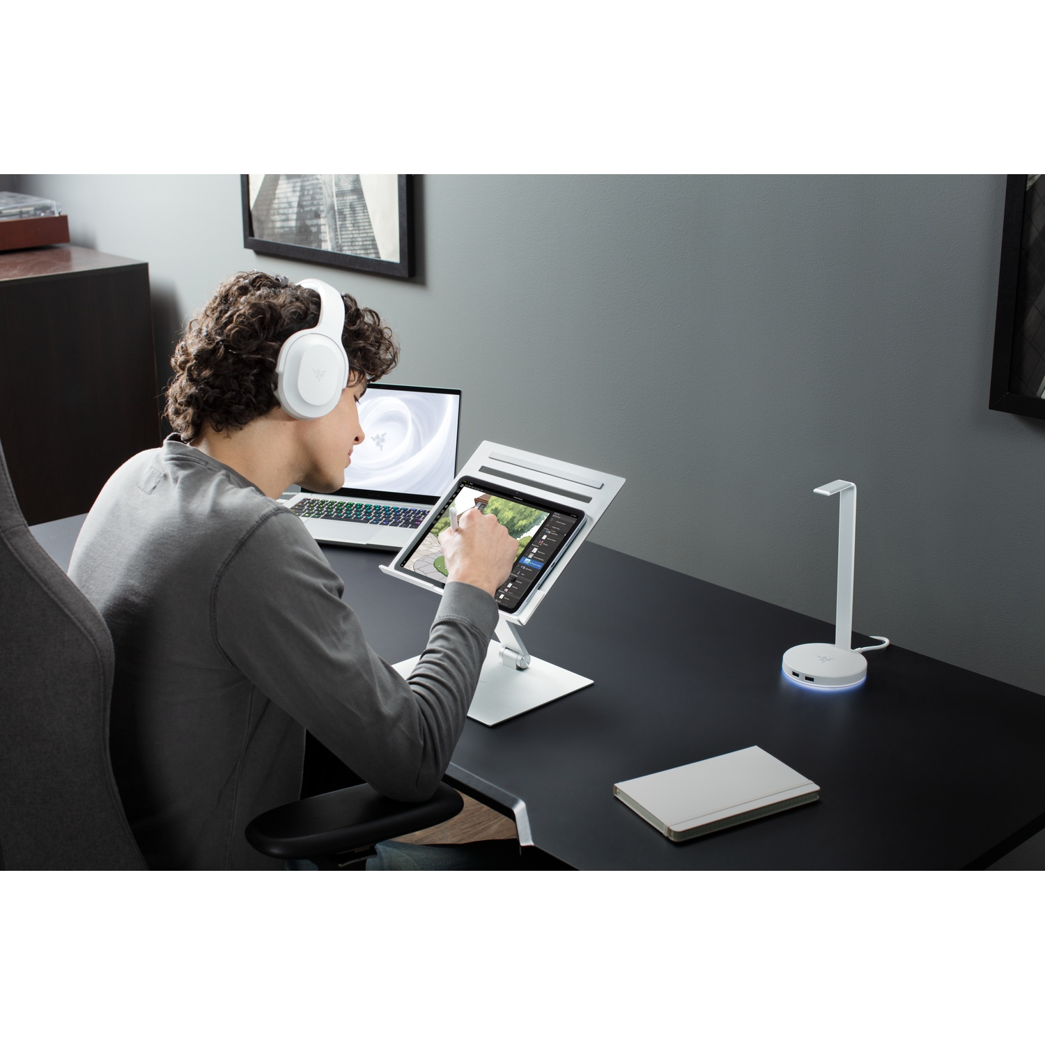 A person wearing headphones is using a tablet on a desk. A laptop, lamp, and notebook are also on the desk.