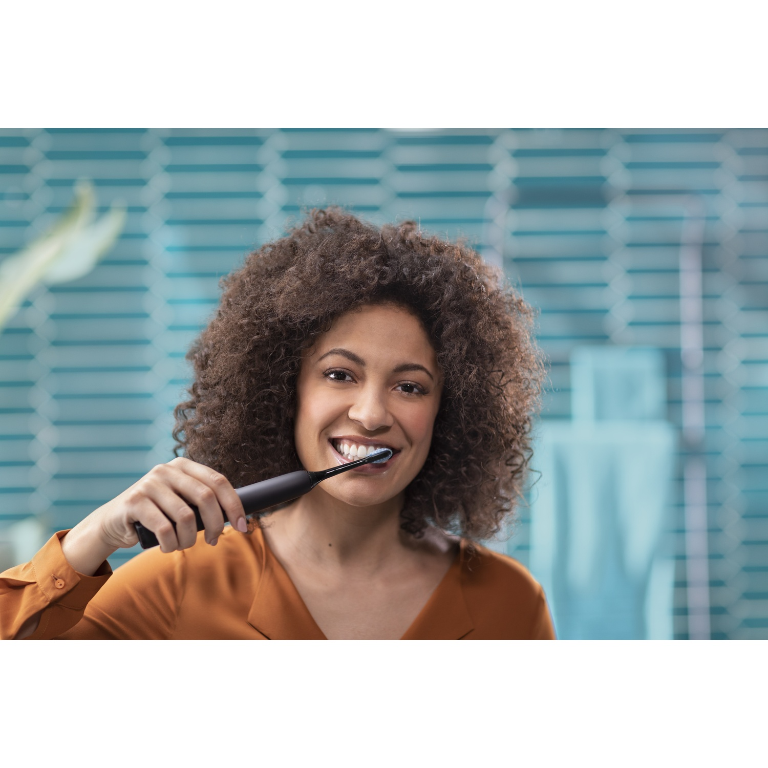 Una donna con i capelli ricci si lava i denti con uno spazzolino elettrico nero, sorridendo. Sfondo: parete blu sfocata.