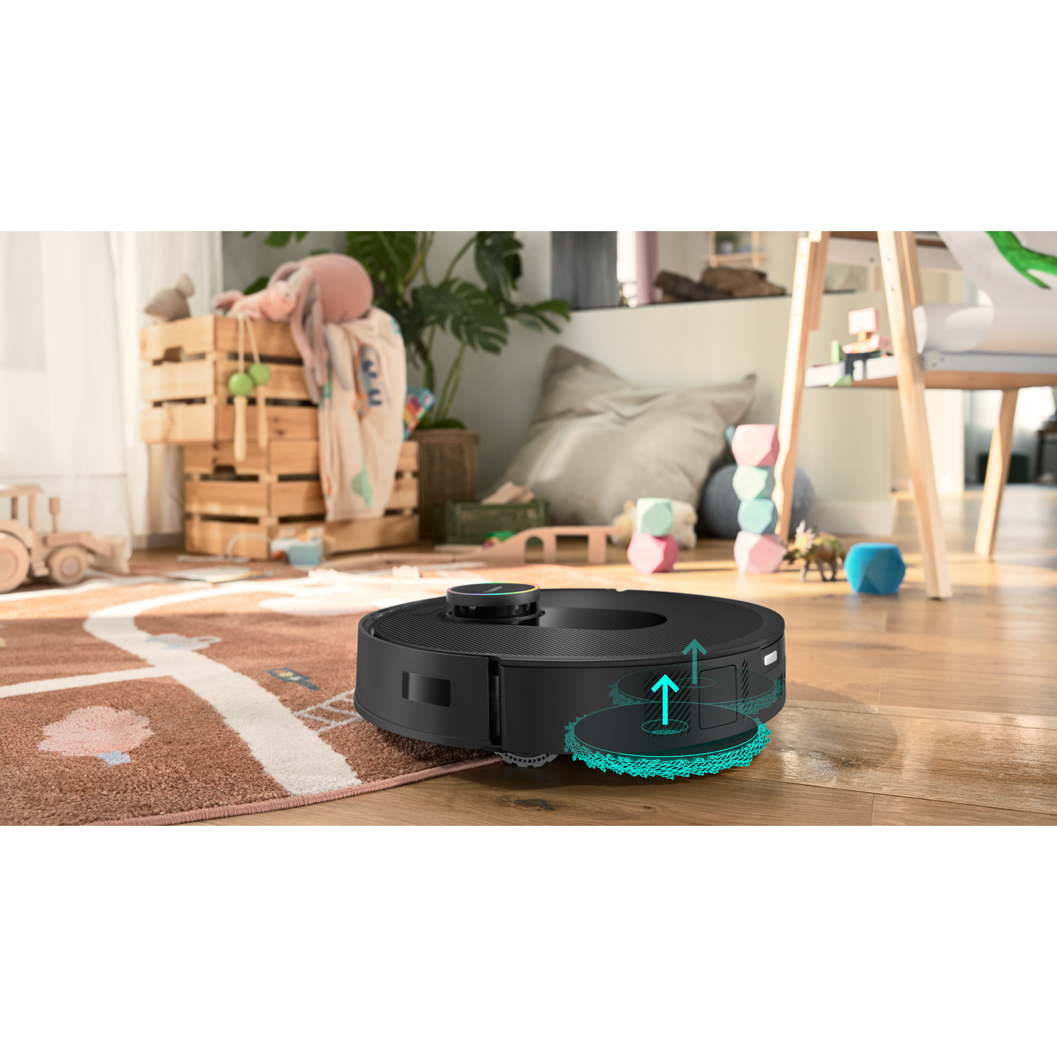 A black robot vacuum cleaner on a wooden floor. Toys and furniture are in the background, including a carpet and wooden boxes.