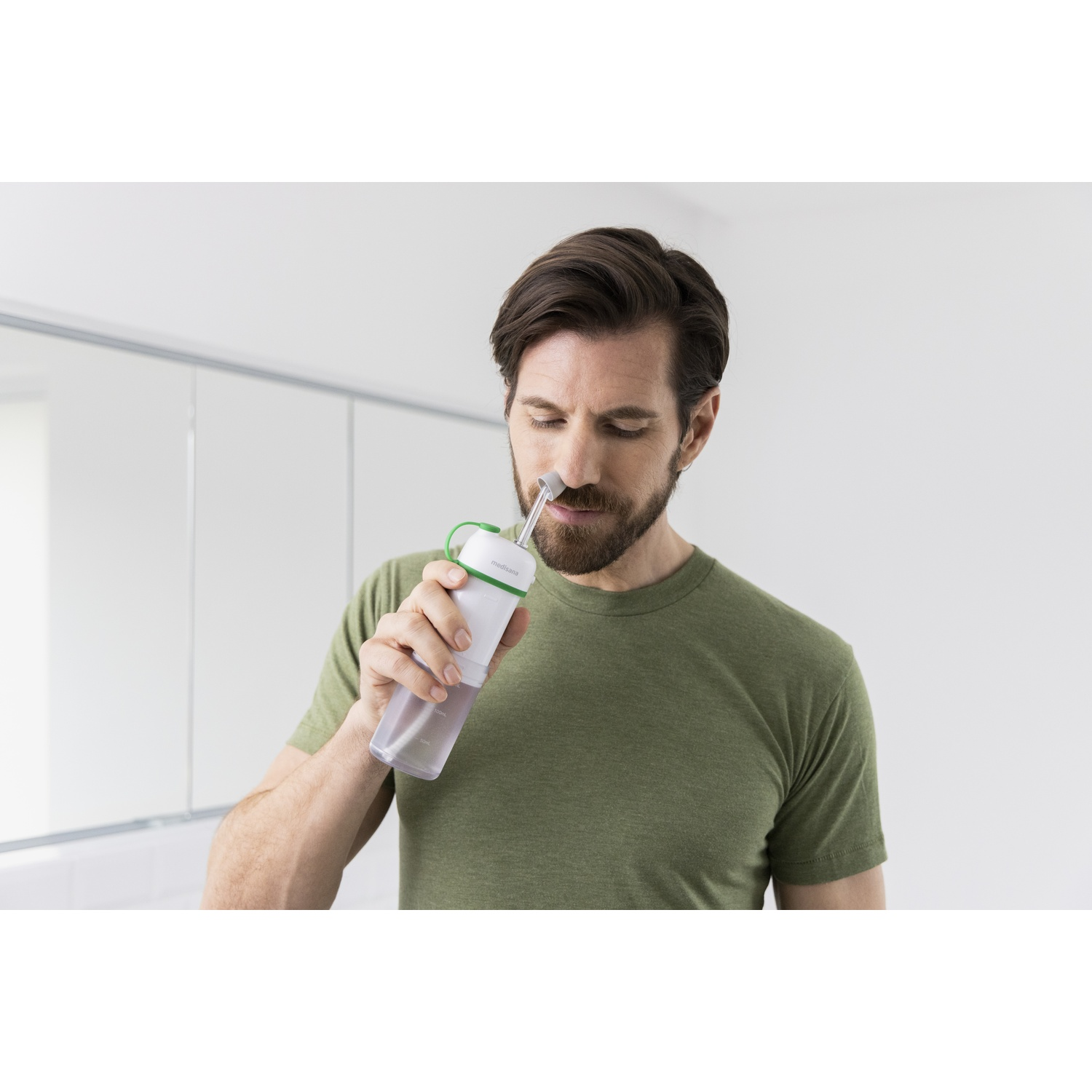 A man is using a nasal rinse bottle. He is wearing a green shirt, looking down, and holding the bottle to his nose.