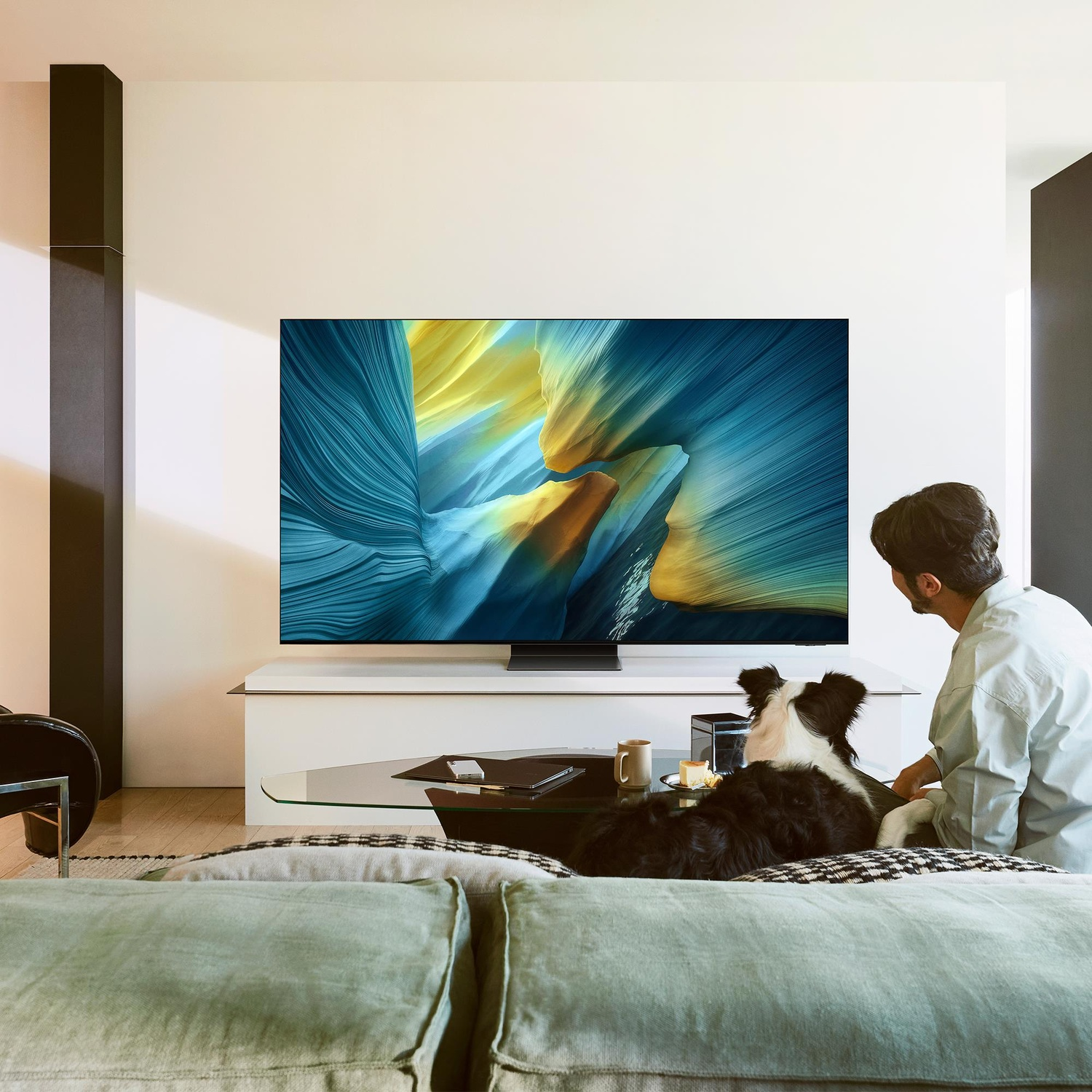 Un uomo e un cane guardano la TV. Un grande schermo mostra arte astratta. Un tavolino ha una tazza e una torta.